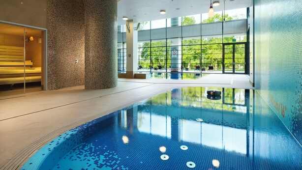 A modern indoor swimming pool with clear glass walls, a bench, and a view of greenery outside. - DoubleTree By Hilton Hotel & Conference Centre Warsaw (Warsaw)