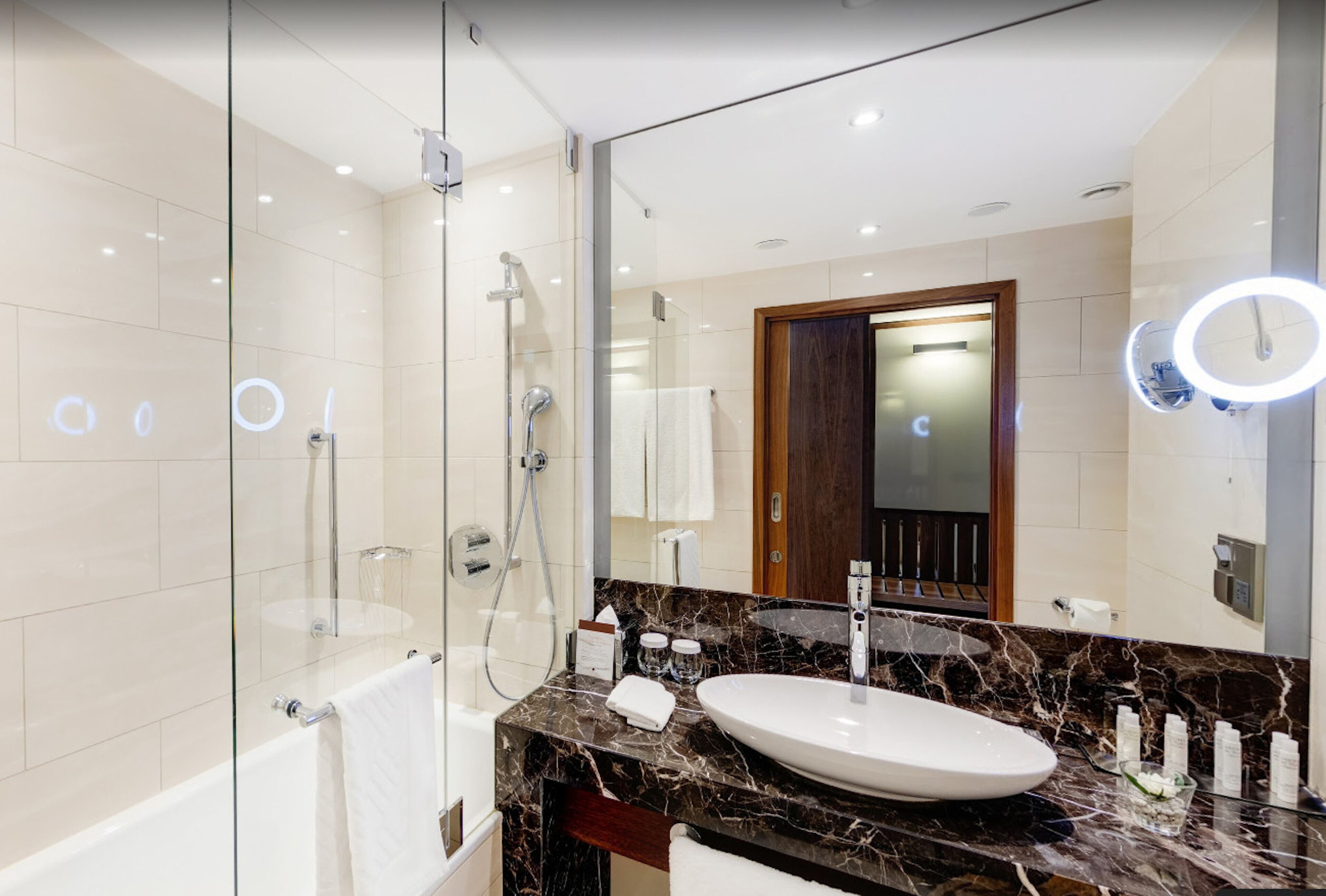 A modern bathroom with a glass-enclosed shower, a white bathtub, a marble countertop, and a large mirror with a circular light.