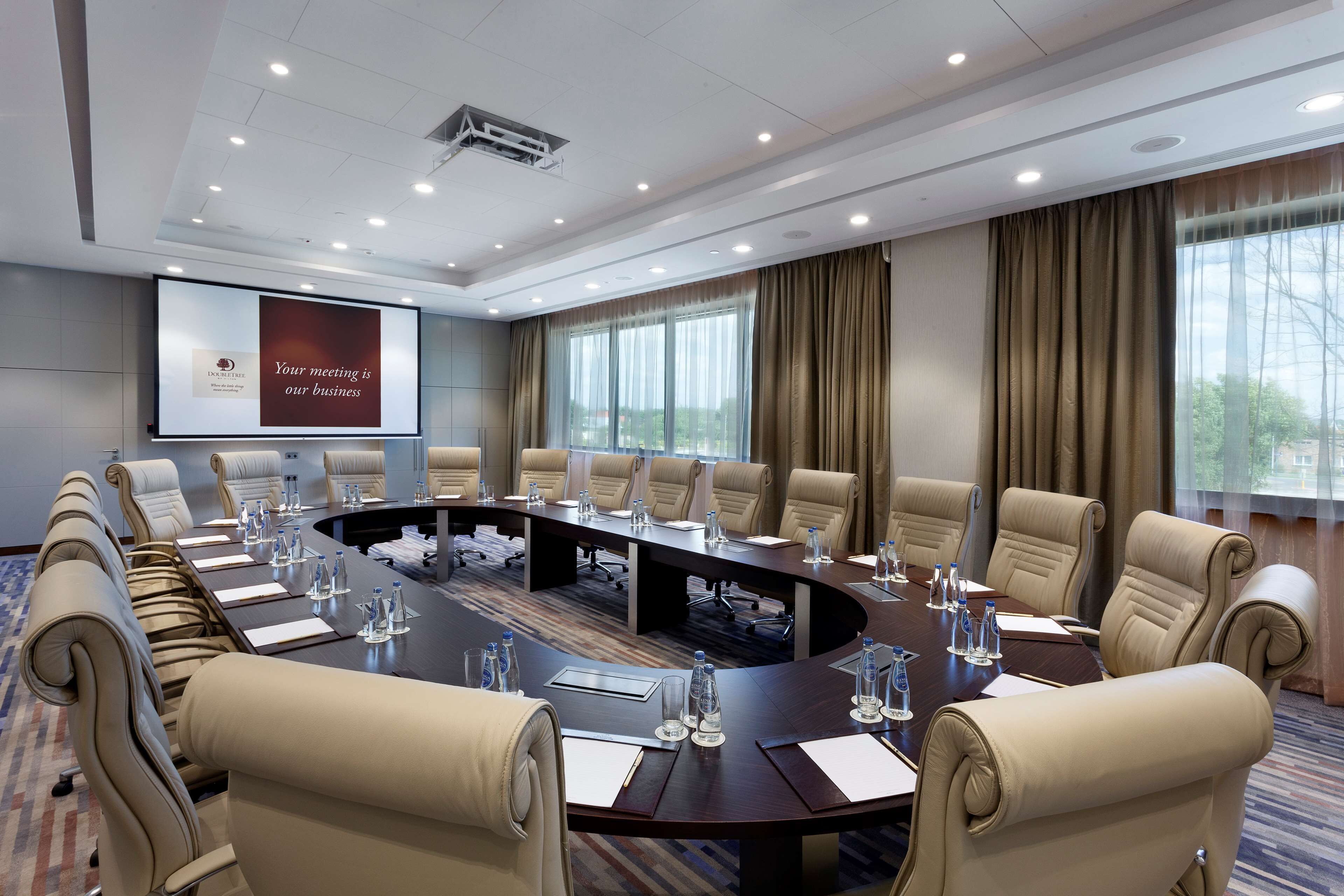 A conference room with a large oval table, beige leather chairs, water bottles, and a projector screen displaying a message.