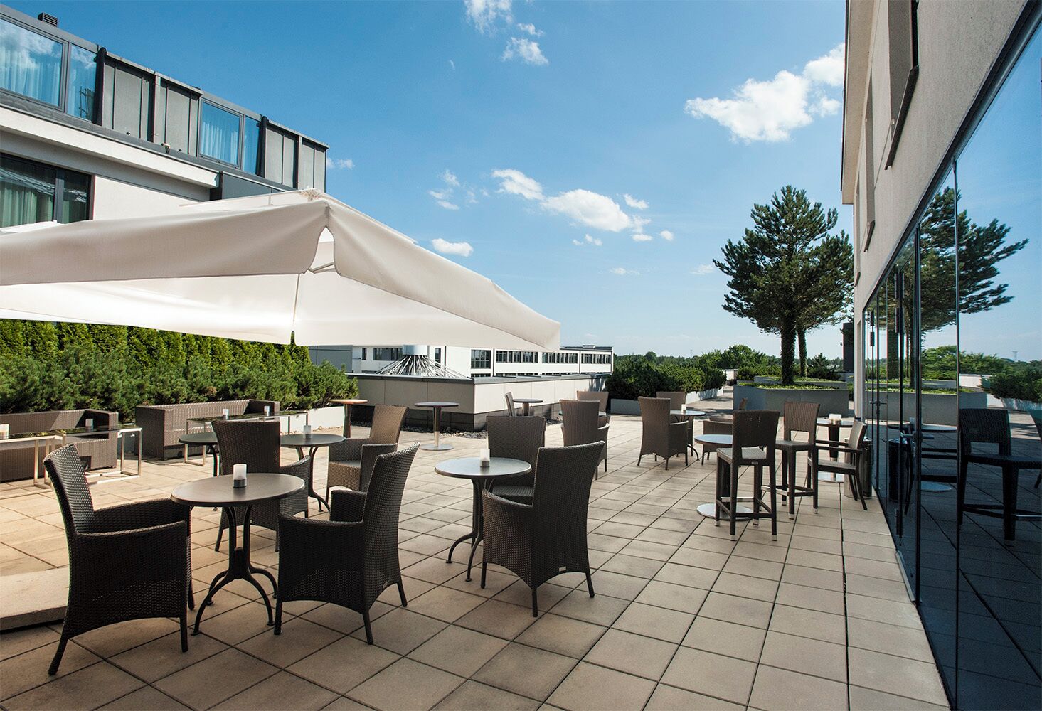 Un patio exterior con mobiliario de mimbre y un amplio paraguas blanco, con vistas a un edificio moderno y un cielo despejado.