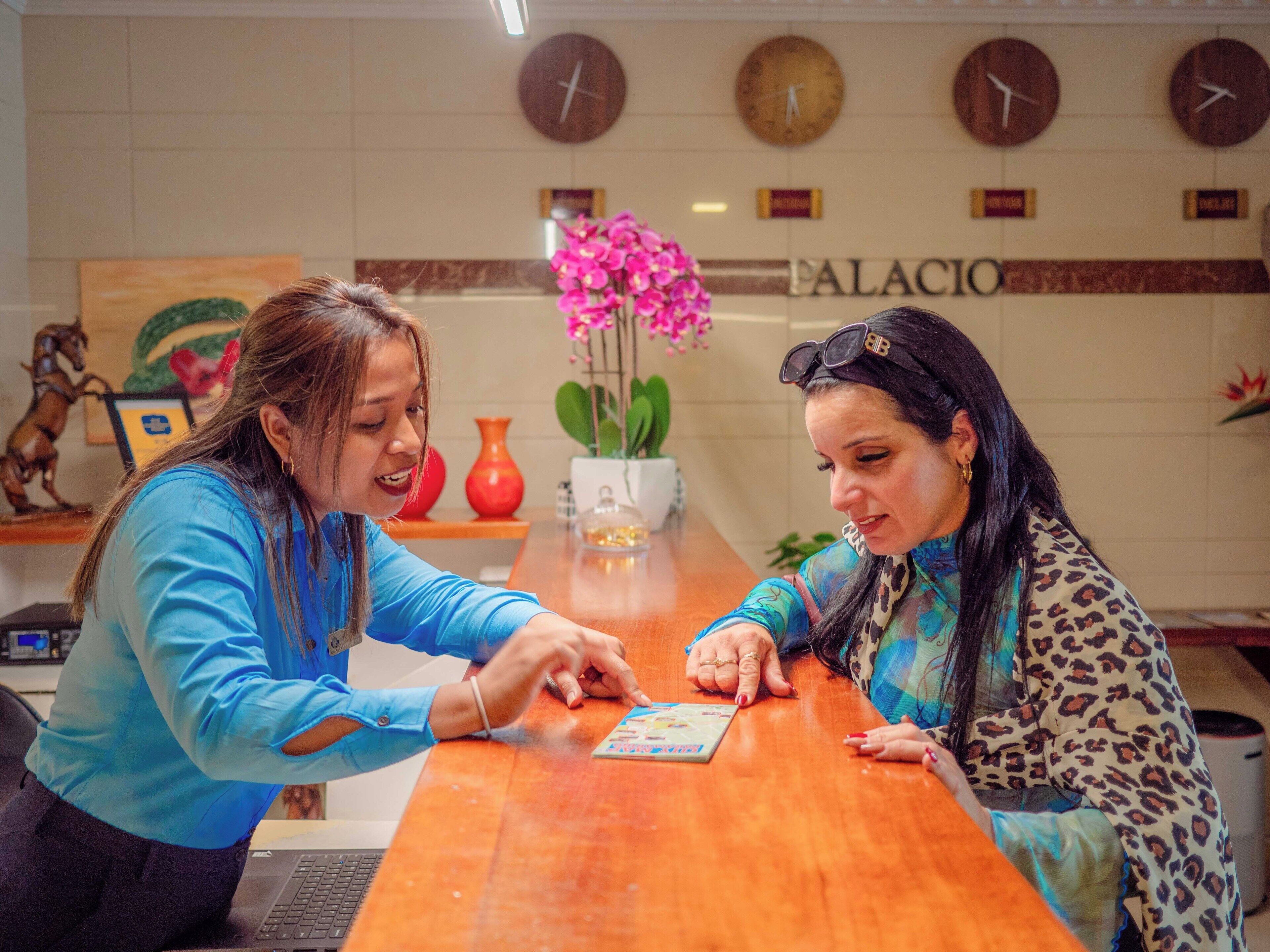 Two people playing a card game at a wooden table in a room with a 'PALACIO' sign on the wall, a clock, and a vase with pink flowers.