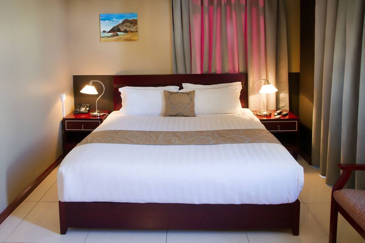 A hotel room with a large bed, two bedside tables with lamps, a painting on the wall, and a view of the beach through curtains.