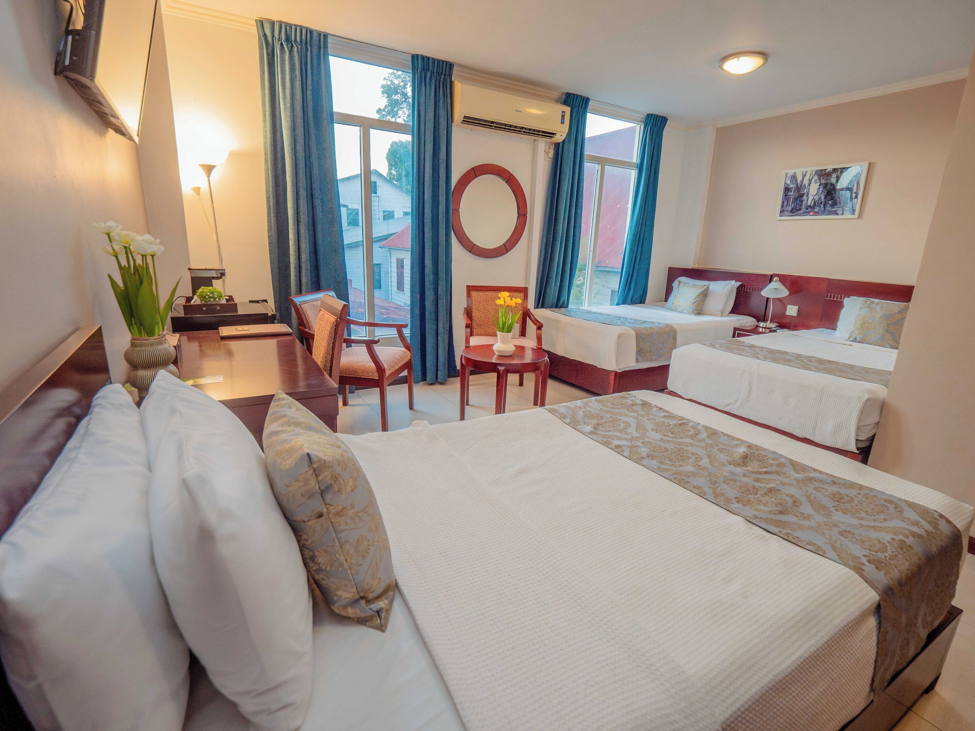 A hotel room with two beds, a desk, chairs, a vase with flowers, and a view of the outside.