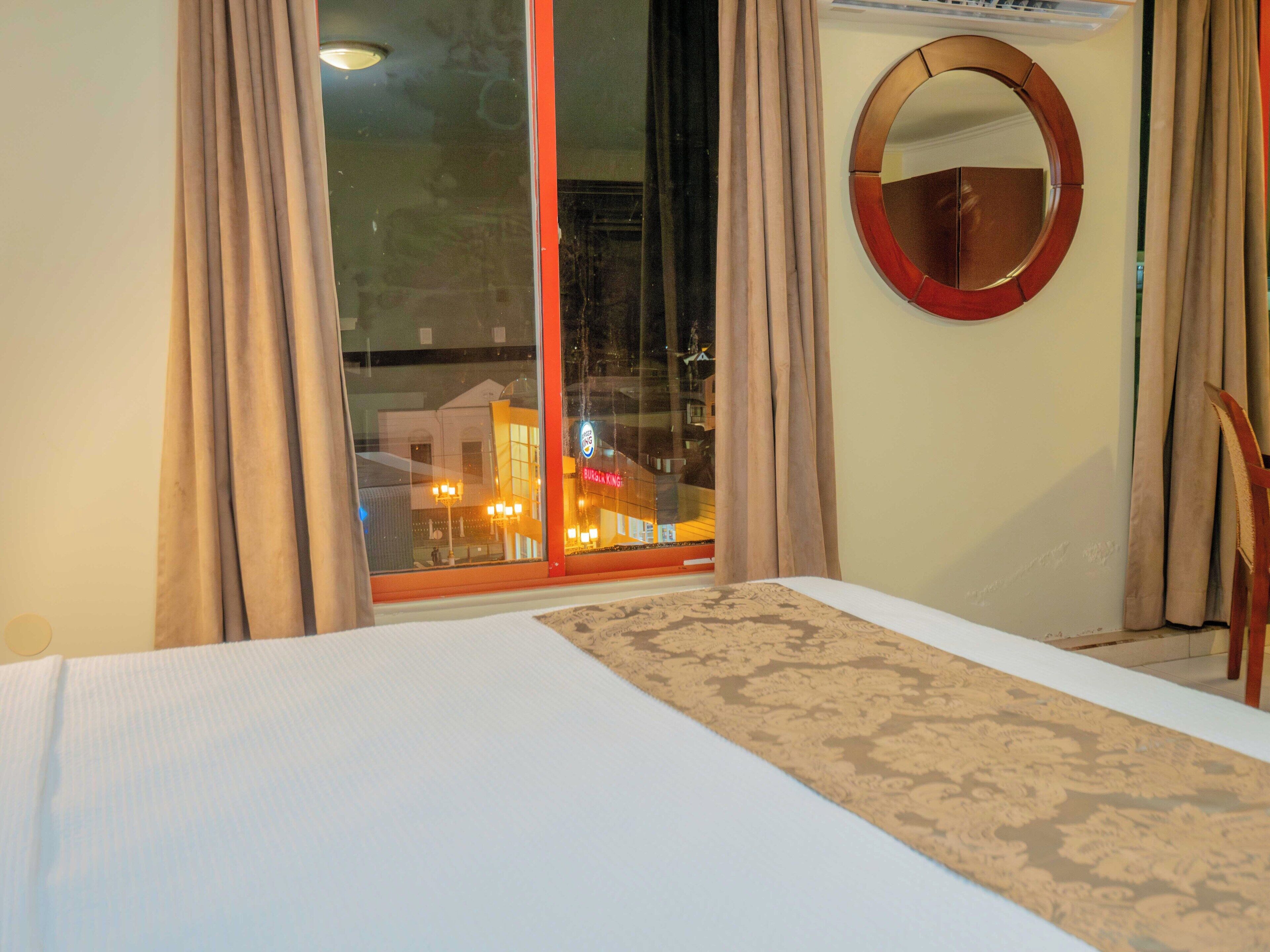 A hotel room with a large bed, a round mirror, and a view of a cityscape through a large window.