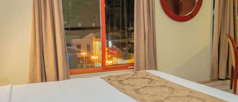 A hotel room with a large bed, a round mirror, and a view of a cityscape through a large window.