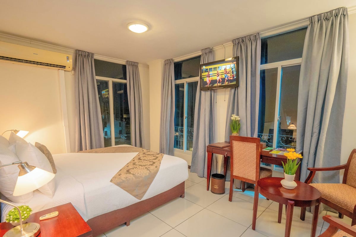 A hotel room with a bed, bedside table, lamp, chair, TV, and a window with curtains.