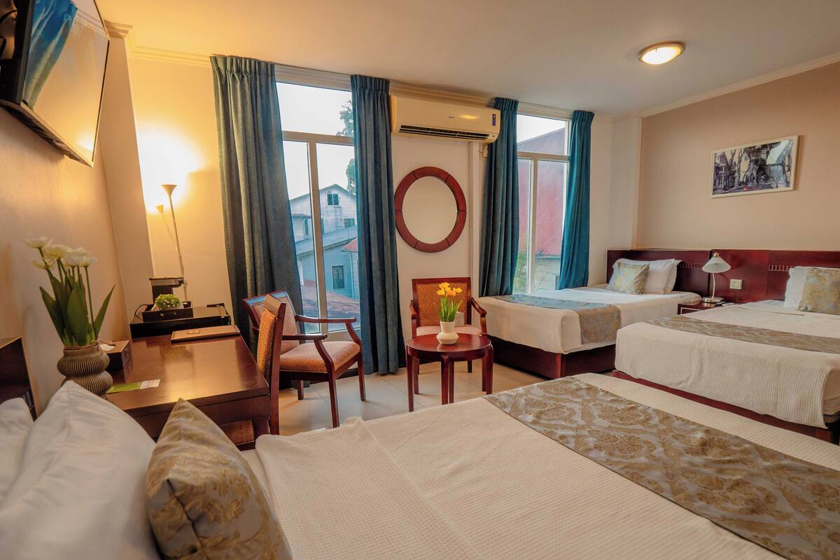 A hotel room with two beds, a desk, a chair, a lamp, a vase with flowers, and a view of the outside.