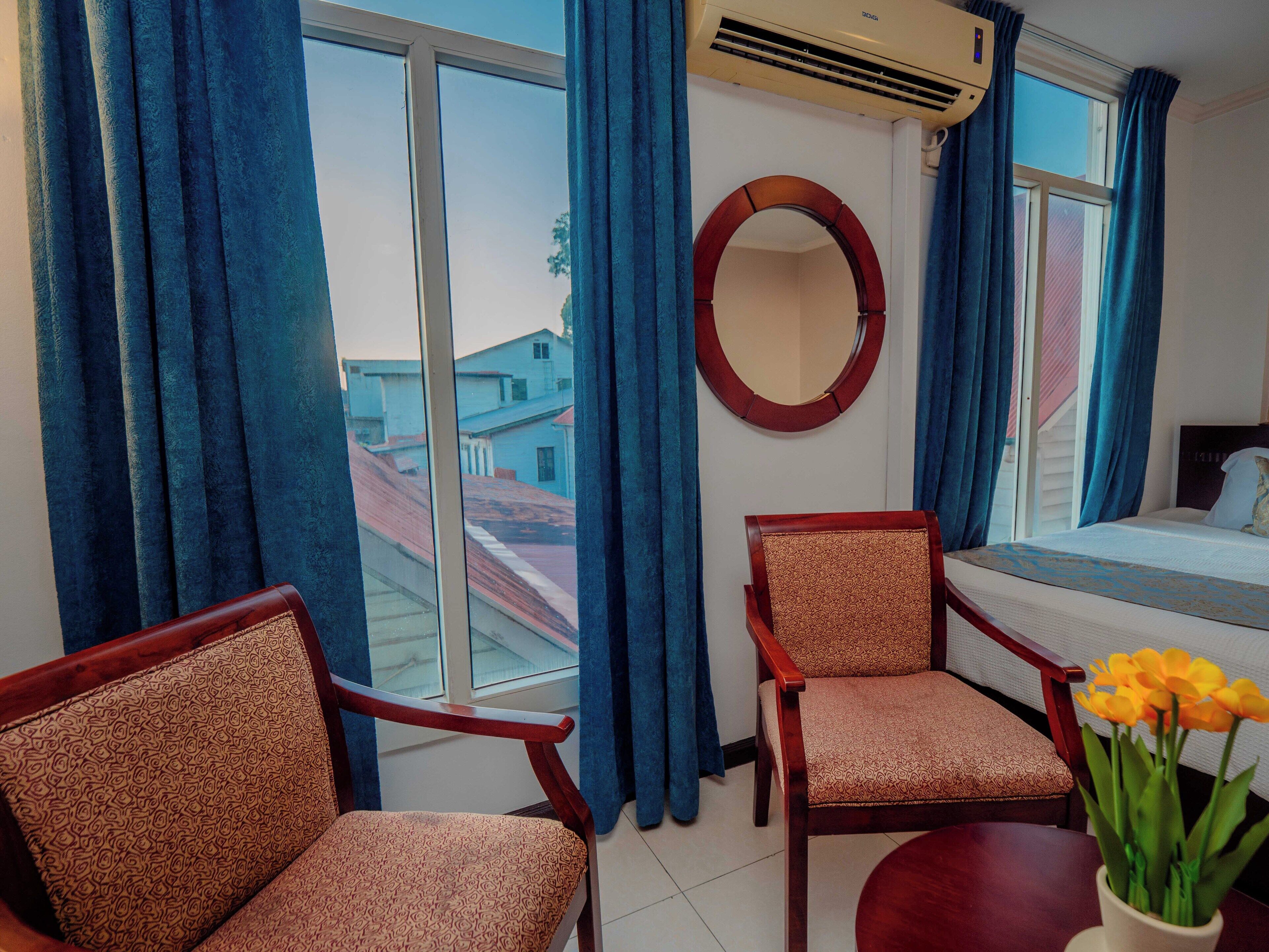 Habitación de hotel con una cama, dos sillas, un espejo redondo y una ventana con cortinas azules.