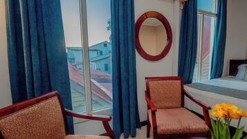 Habitación de hotel con una cama, dos sillas, un espejo redondo y una ventana con cortinas azules.
