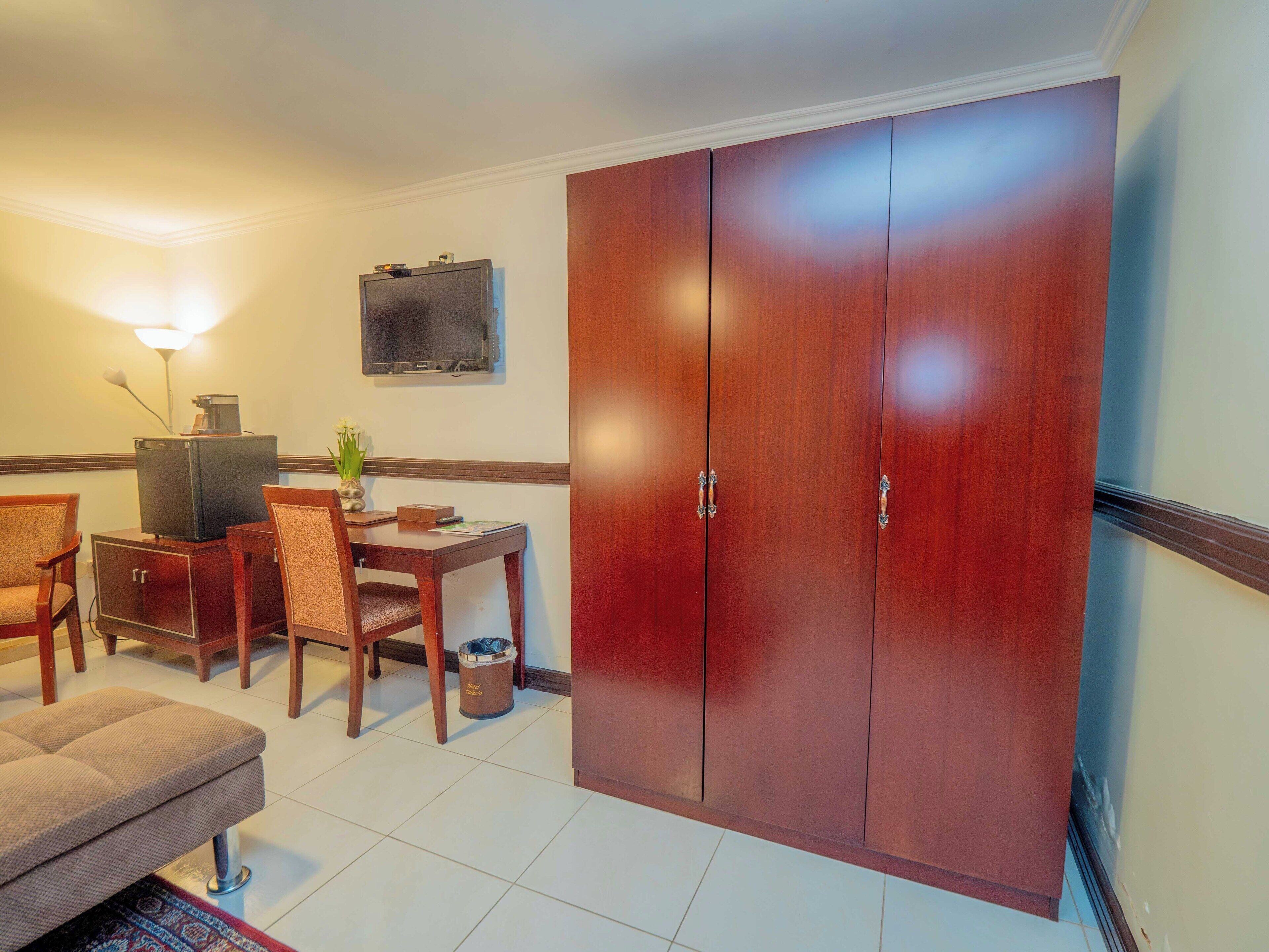 A hotel room with a wooden wardrobe, a small dining table with chairs, a television mounted on the wall, and a lamp.