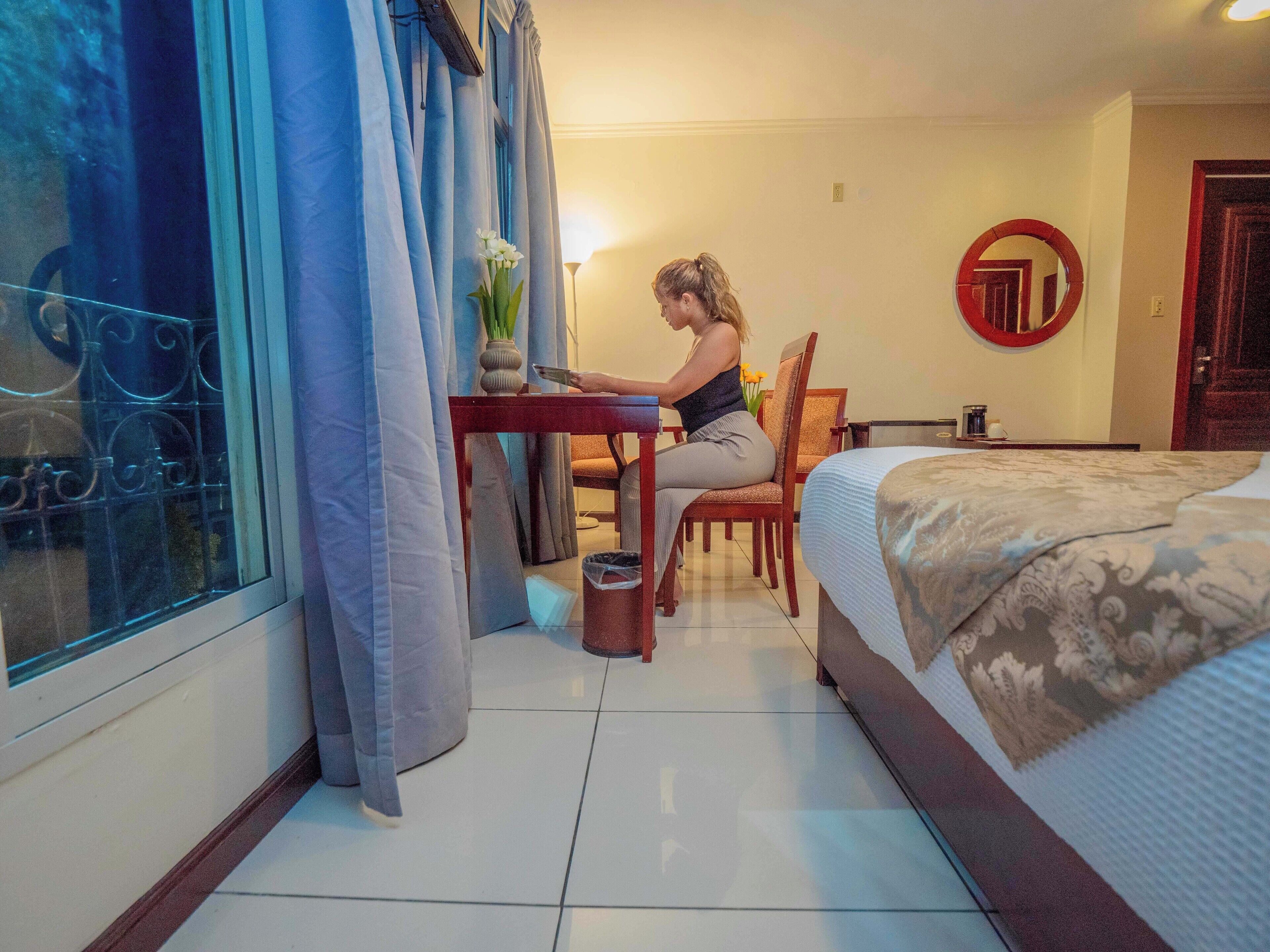 Habitación de hotel con cama, escritorio y silla. Hay una ventana con cortinas, un espejo en la pared y un florero con flores en el escritorio.