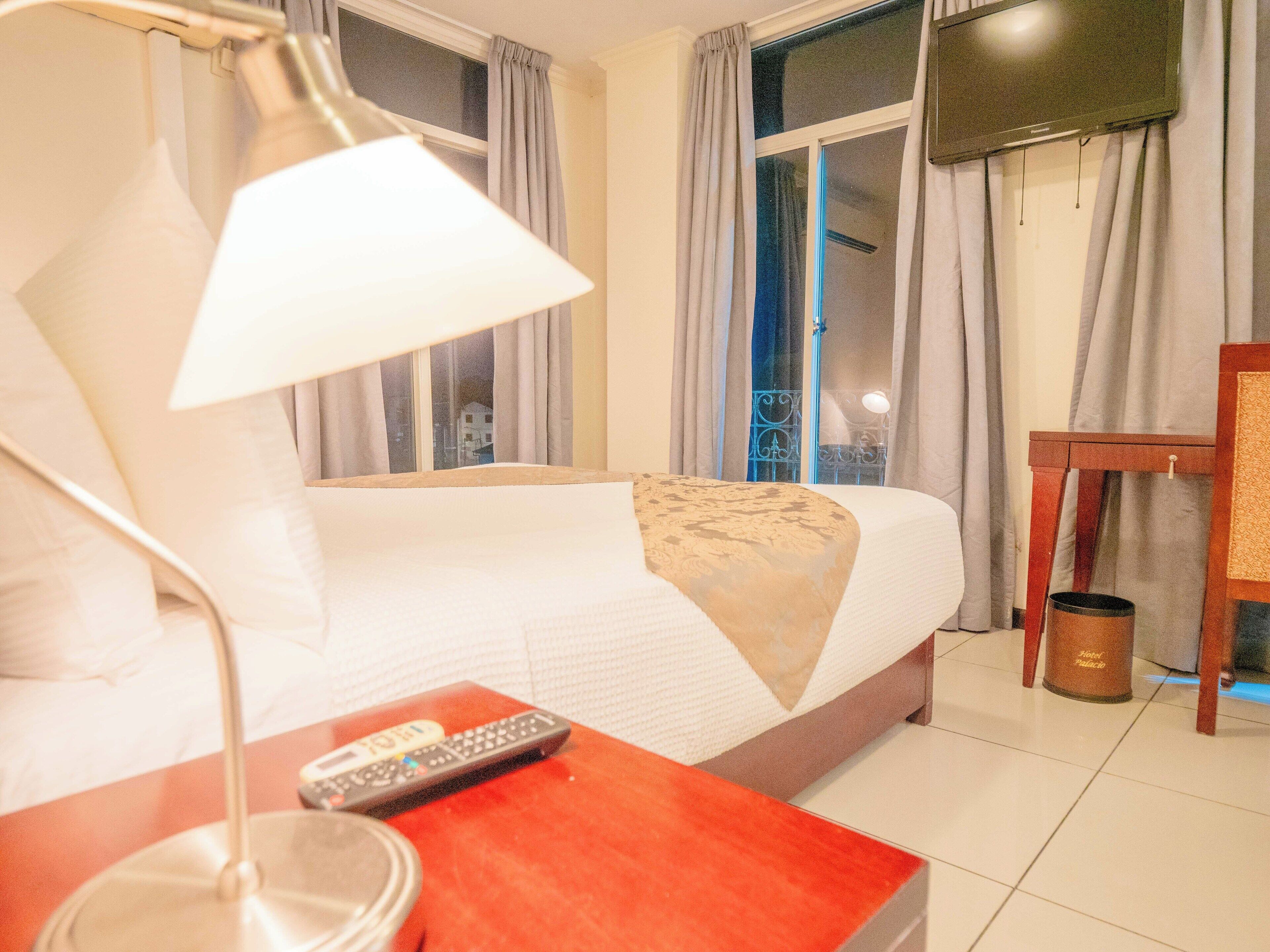 A hotel room with a bed, a lamp, a remote control, a TV, and a trash bin.