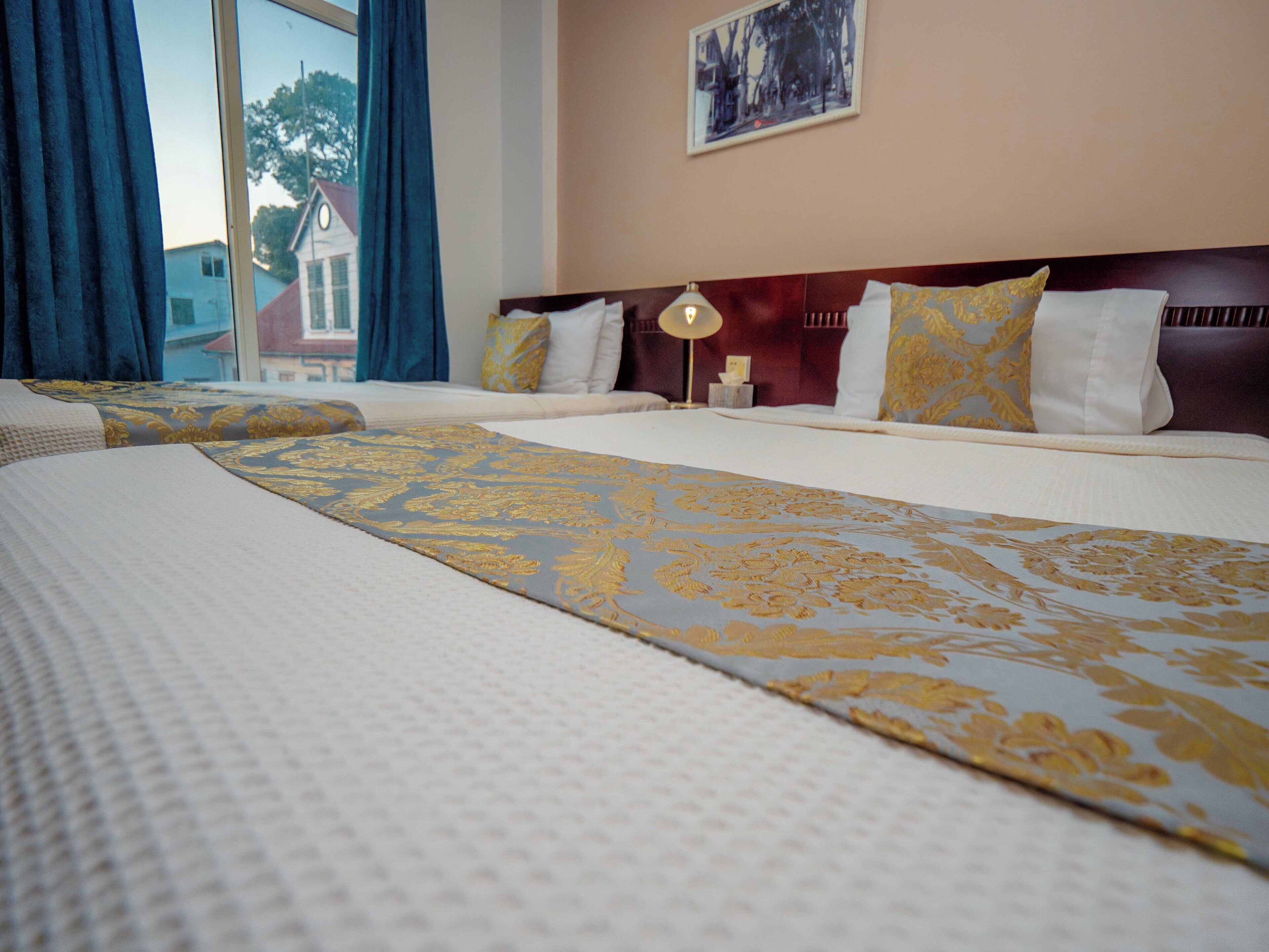 A hotel room with two beds, a large window with curtains, a framed picture on the wall, and a bedside lamp.