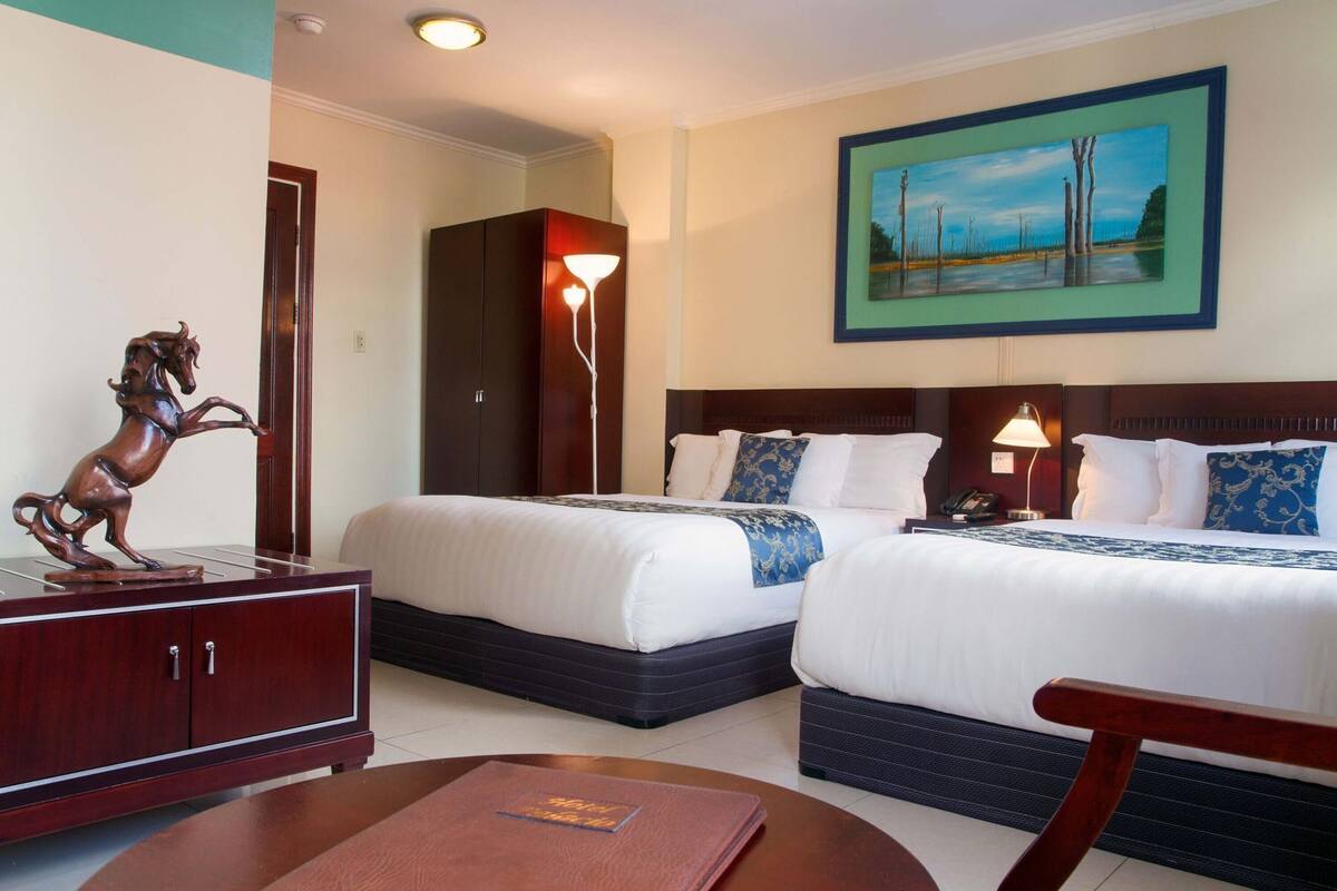 A hotel room with two beds, a wooden cabinet with a horse sculpture, a painting of trees, and a nightstand with a lamp.