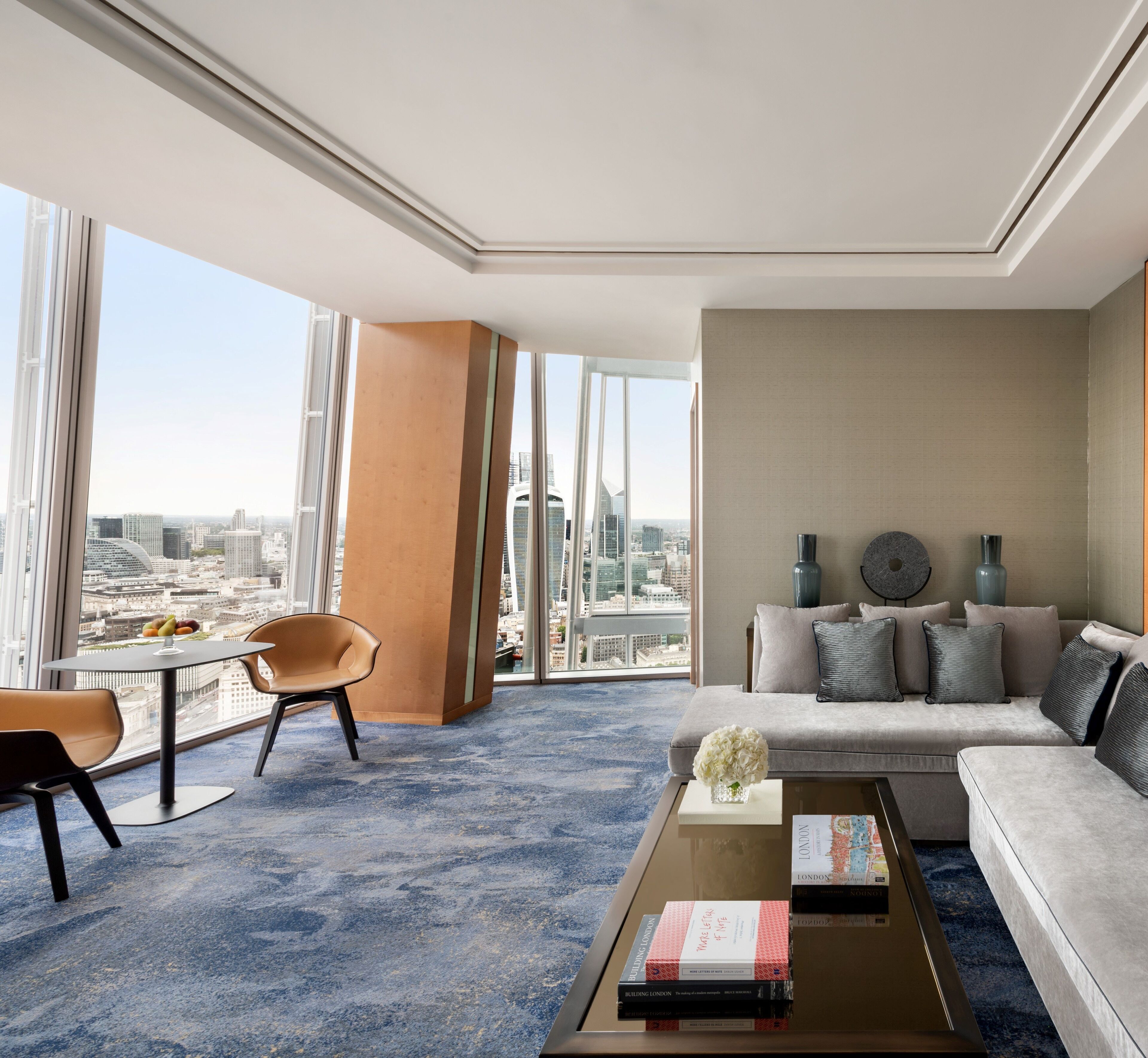 Une chambre d’hôtel moderne dotée d’une grande fenêtre donnant sur la ville, d’un ensemble de canapés, d’une table basse avec des livres et de deux fauteuils.