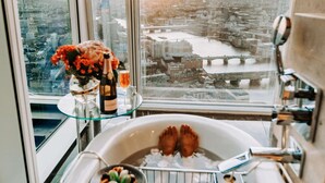 A high-rise room with a large window overlooking a cityscape, a bathtub, a glass table with a bottle and flowers, and a person reading a book. - Shangri-La The Shard, London (London)