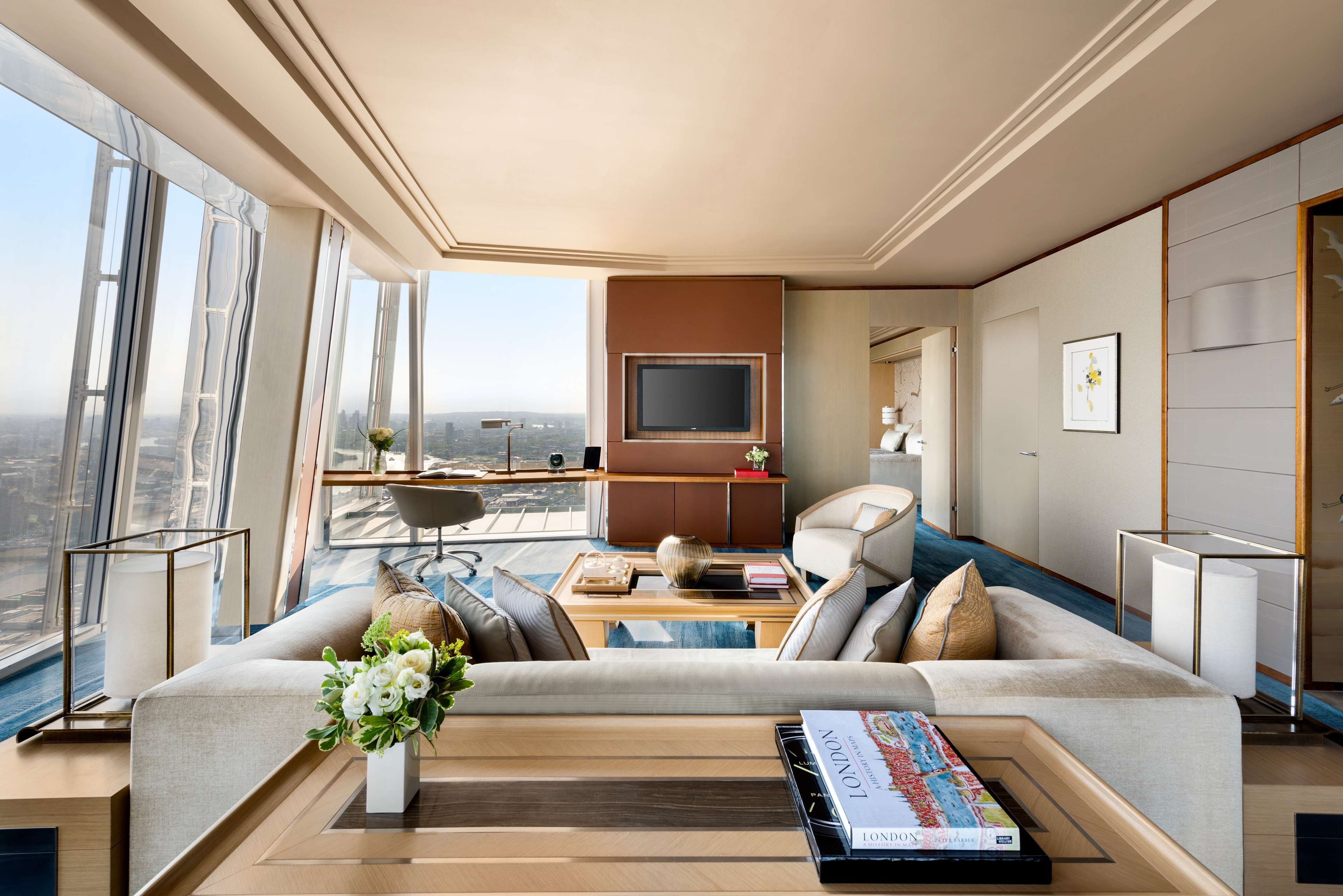 A spacious living room with a large sofa, a coffee table, and a flat-screen TV. There is a balcony with a view of the city.