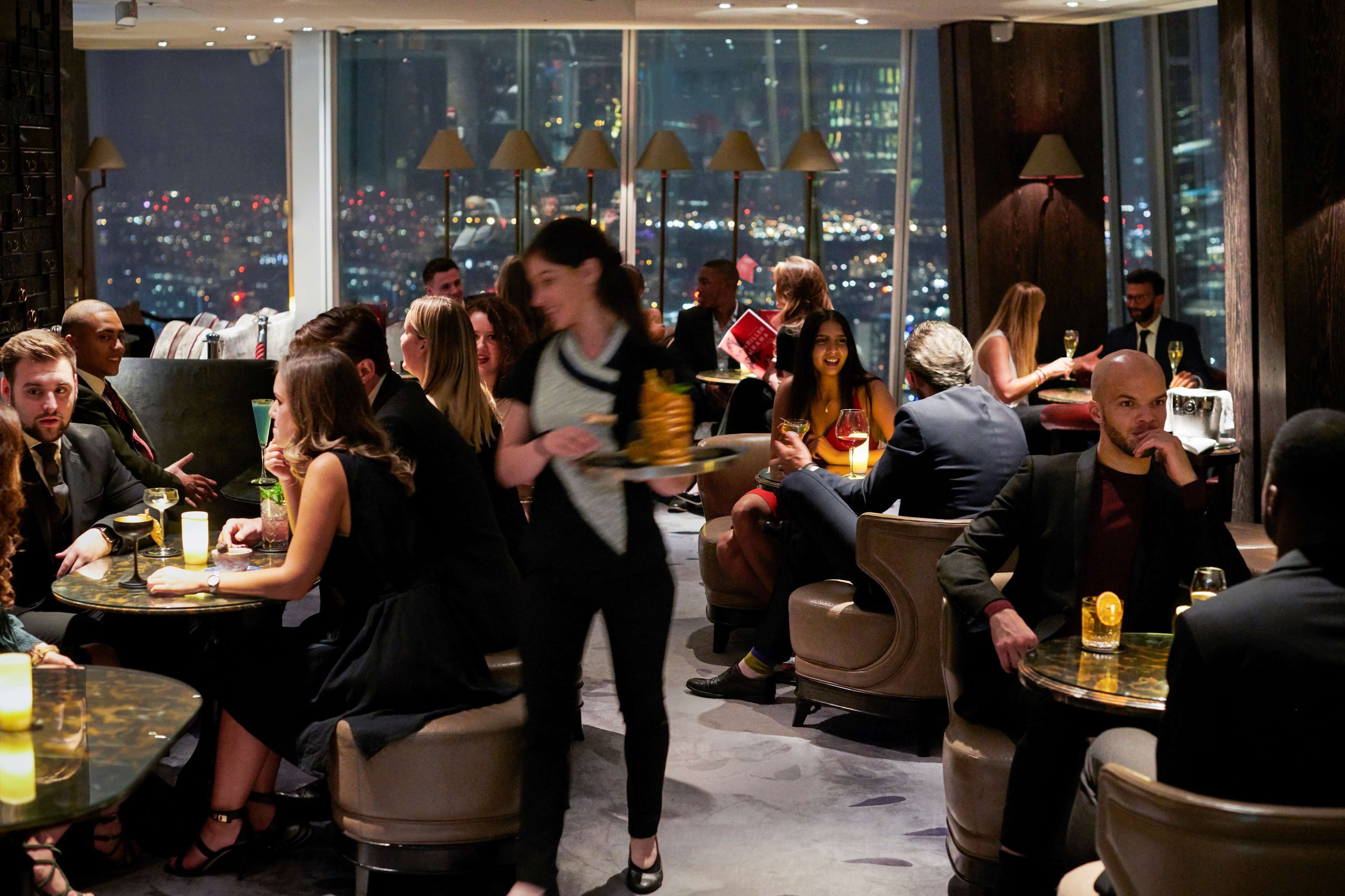 A bustling indoor dining area with round tables and chairs, large windows offering a city view, and ambient lighting.