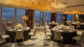 Un ristorante di un grattacielo con tavoli rotondi apparecchiati per la cena, ampie finestre che offrono vista sulla città e sontuose lampadari.