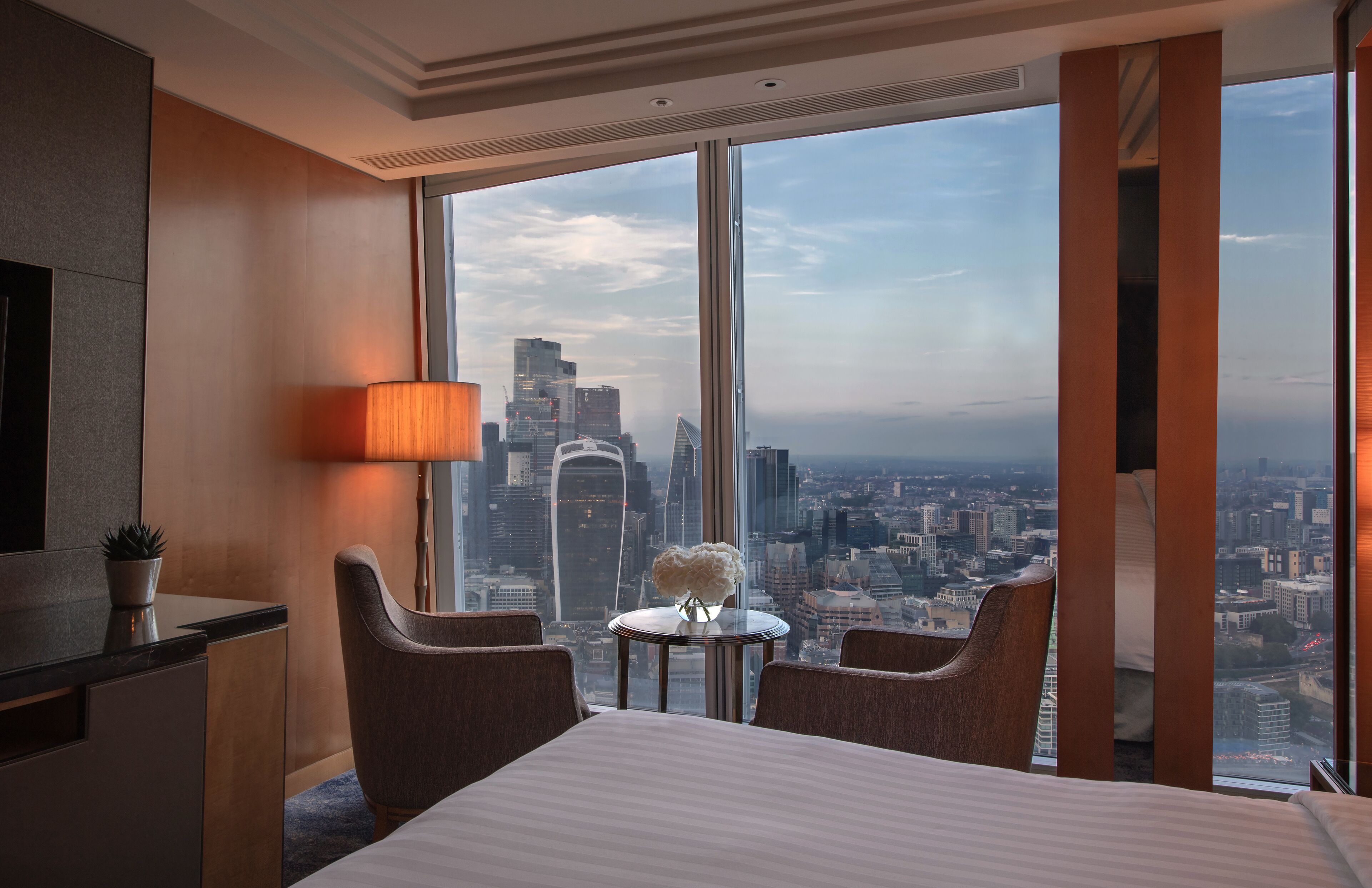 A hotel room with a large window offering a city view, a bed, two chairs, a small table, and a lamp.