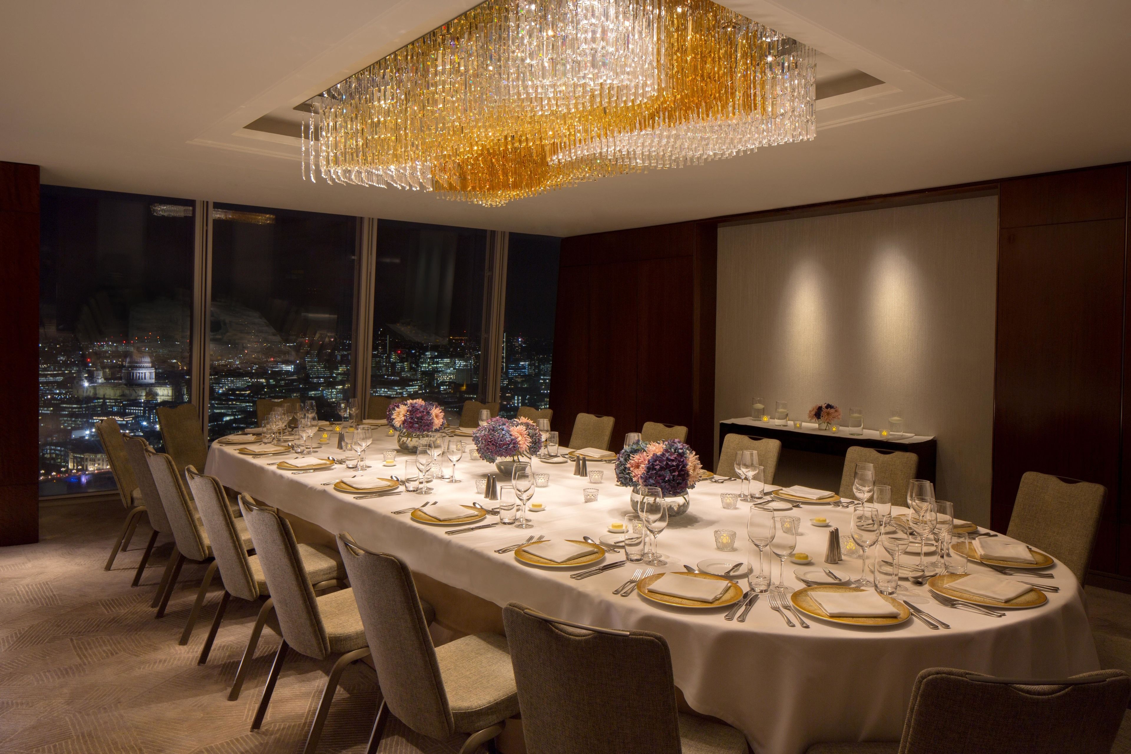 A dining room with a long table set for a meal, large windows, and a city view at night.