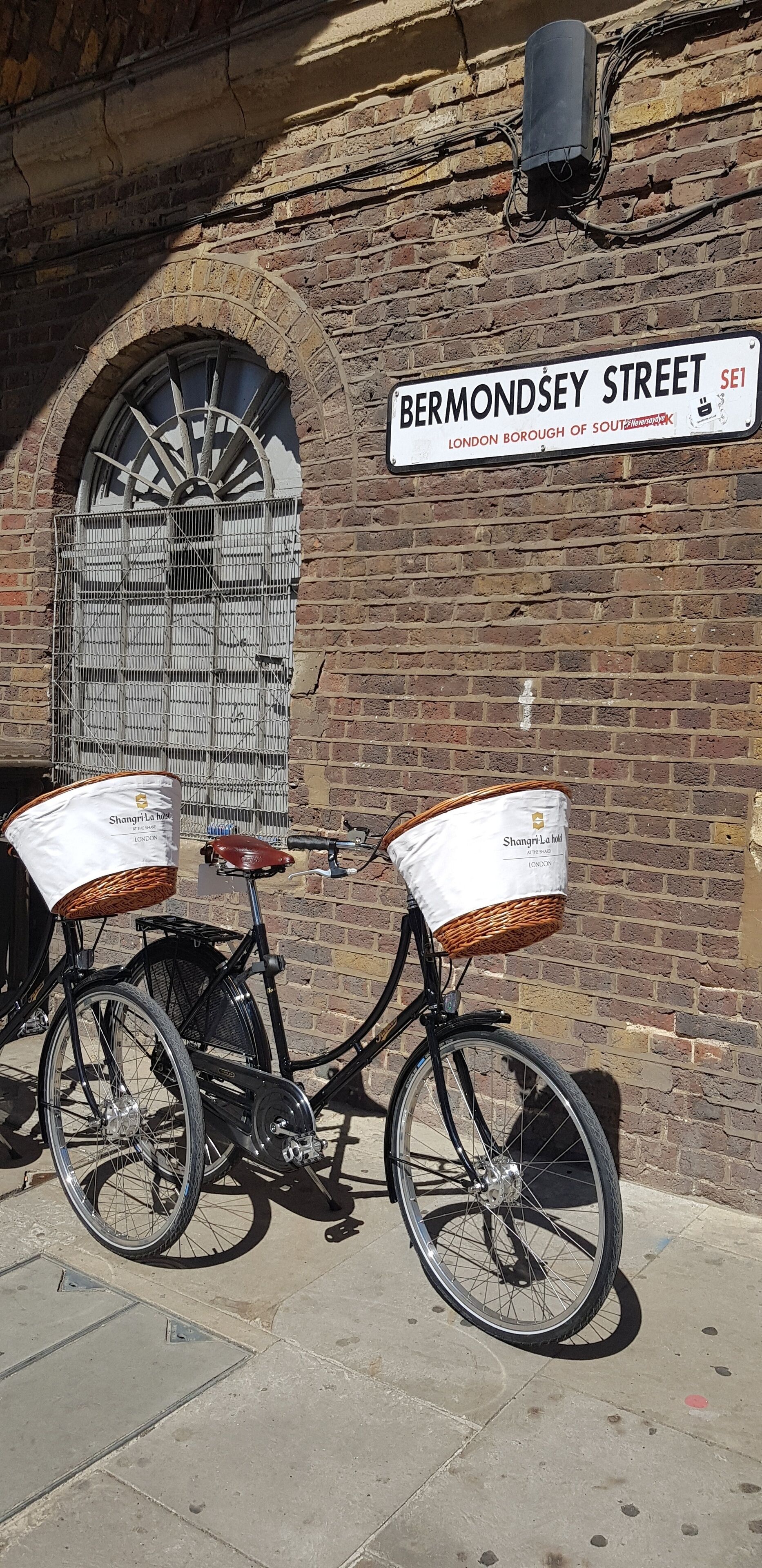 A bicycle with baskets attached to the front and back, parked against a brick wall with a street sign reading "BERMONDSEY STREET" and "LONDON BOROUGH OF SOUTHERNWARK."
