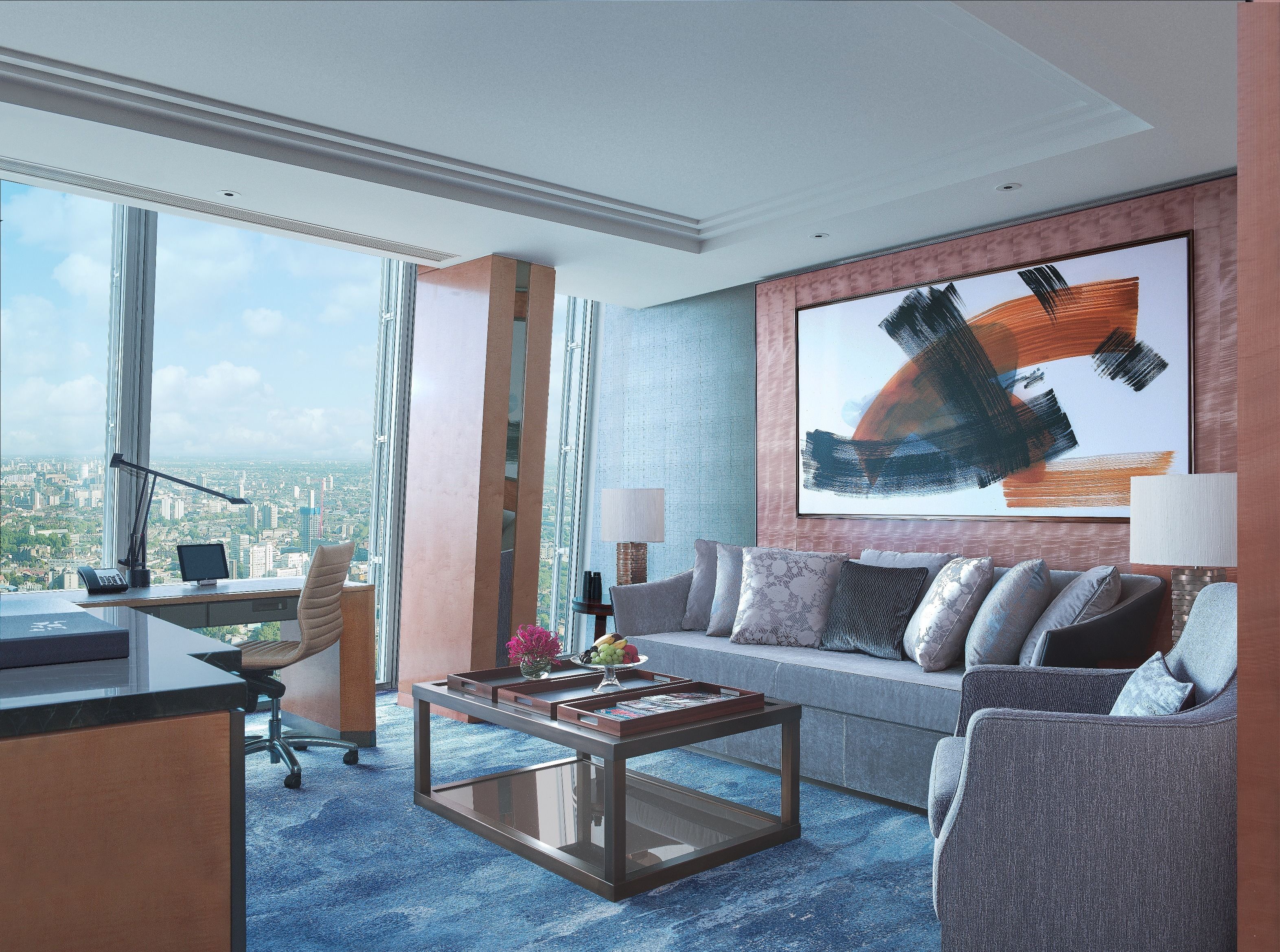 A modern hotel room with a city view, a large abstract painting, a sofa, a coffee table, and a desk with a computer.