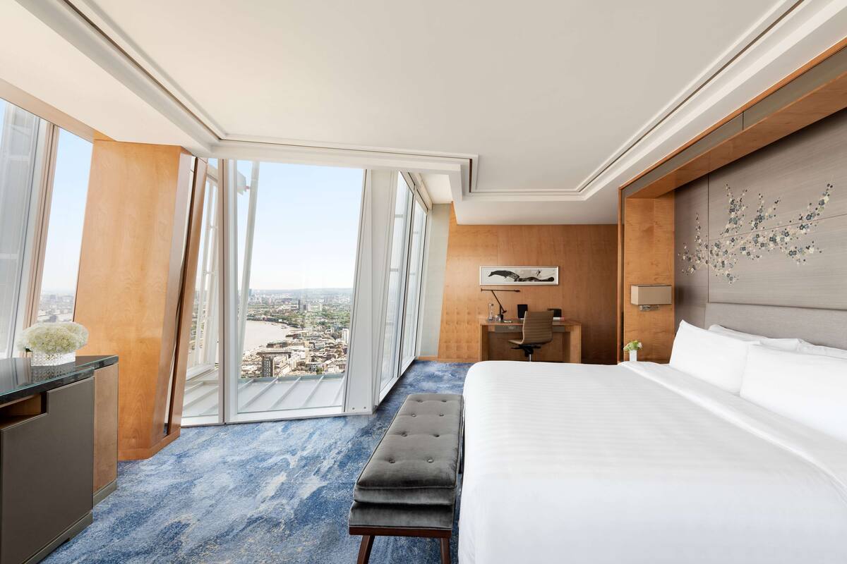 A modern hotel room with a large bed, a desk, and a view of the city through floor-to-ceiling windows.