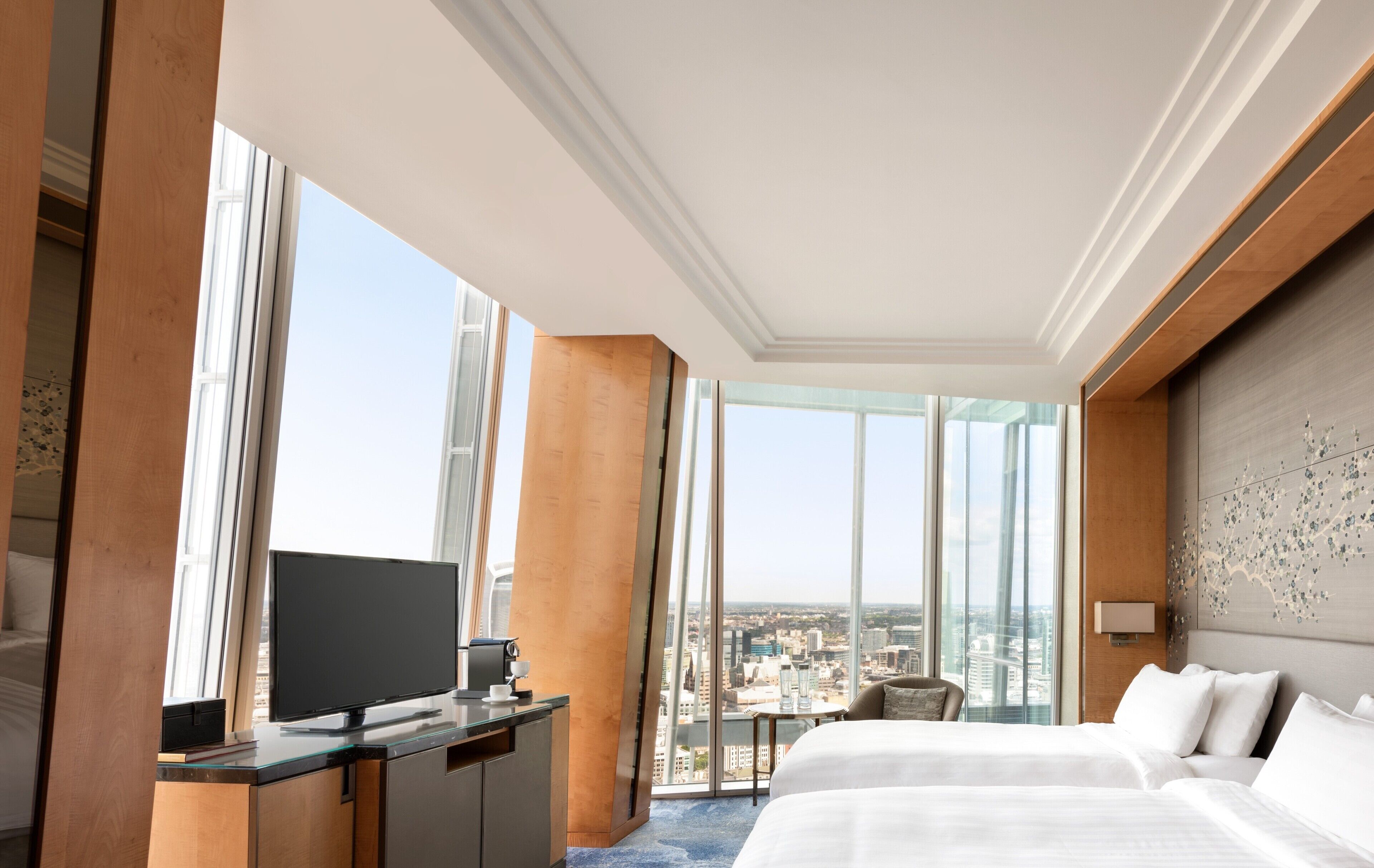 A hotel room with a large bed, a desk with a TV, and a city view through large windows.