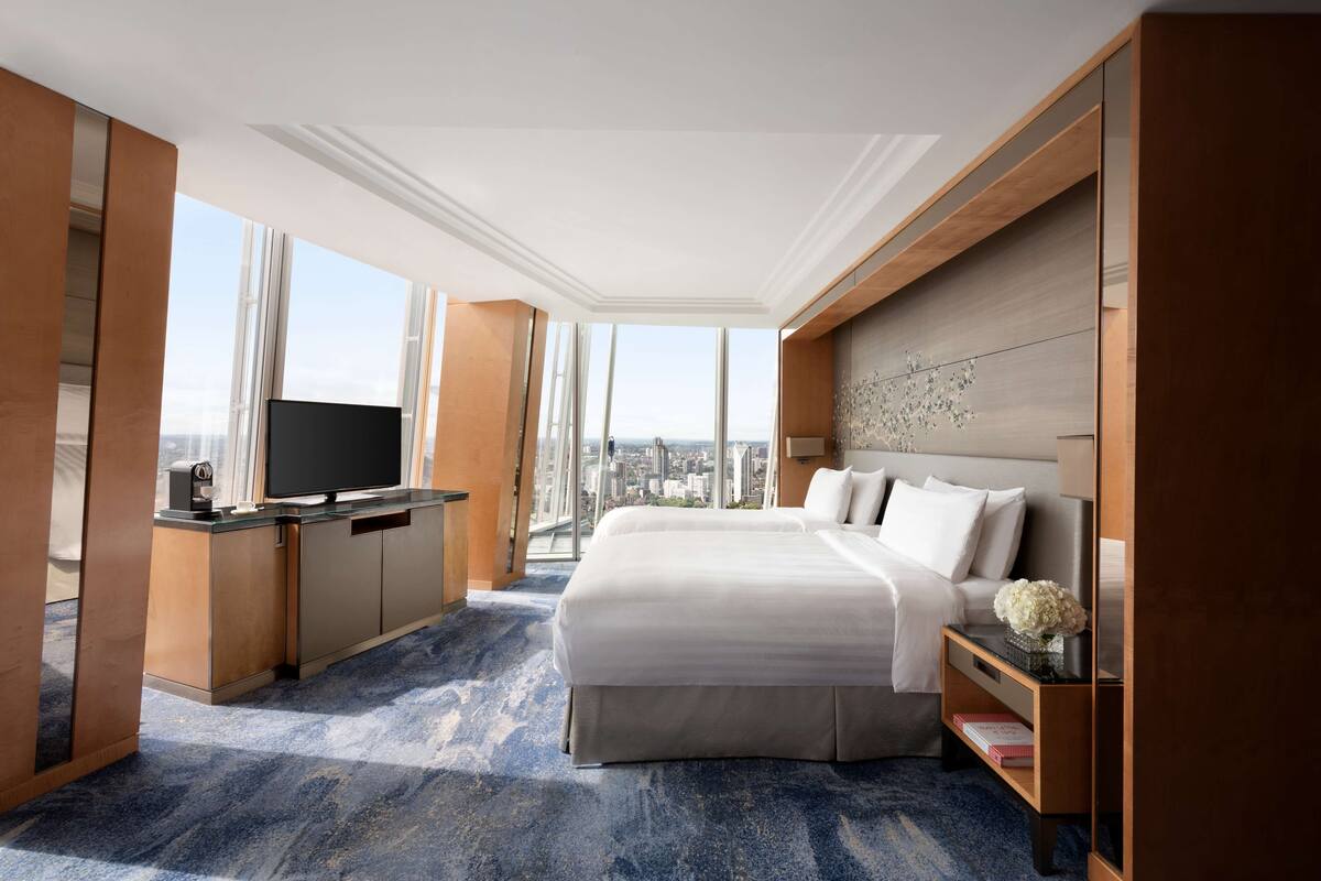 A hotel room with a large bed, a desk with a flat-screen TV, and a view of the city through large windows.