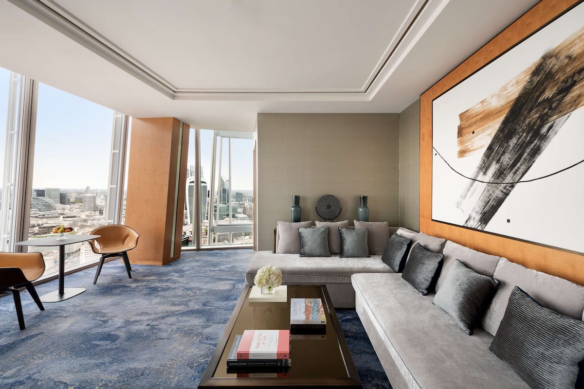 A modern hotel room with a large window offering a city view, a grey sofa, a coffee table with books, and a round table with chairs.