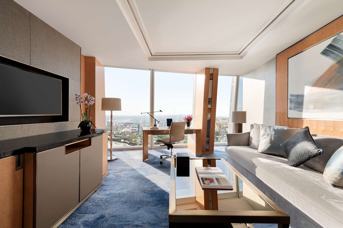 A modern hotel room with a large TV, a sofa, a desk, and a city view.