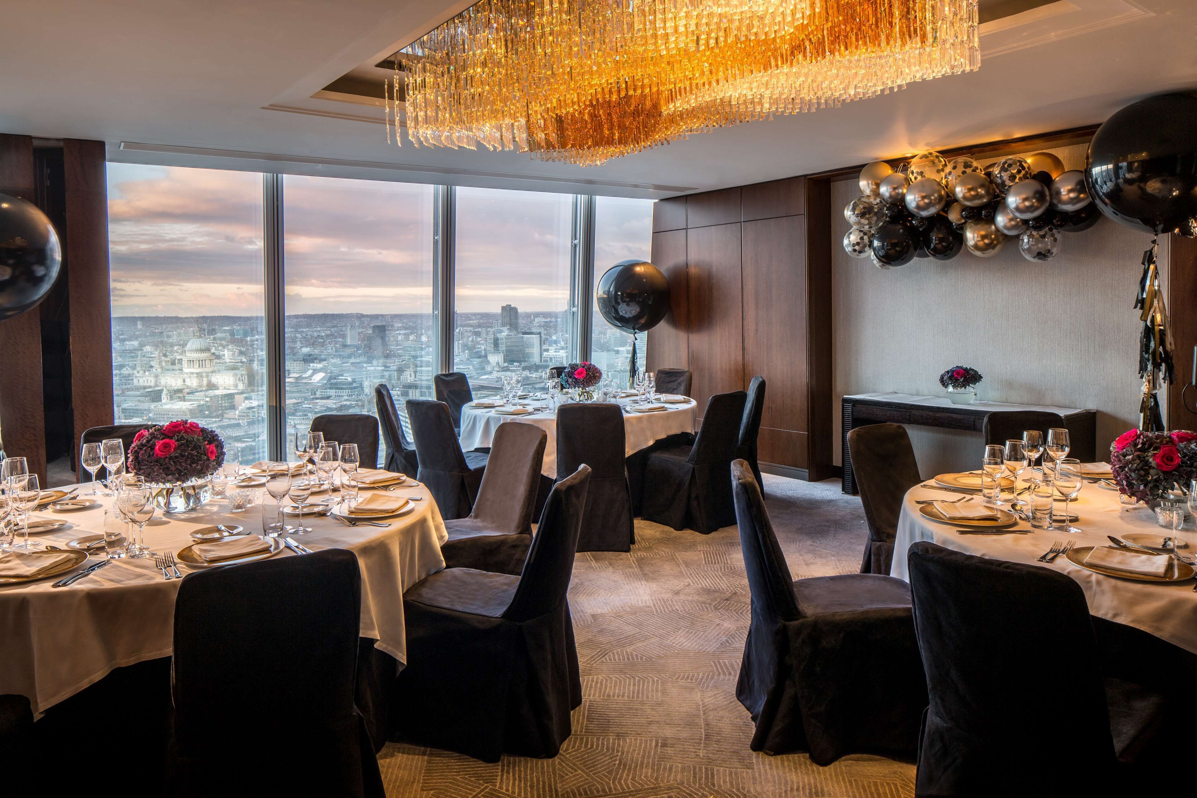 A dining area with tables set for a meal, large windows with a city view, and a chandelier.