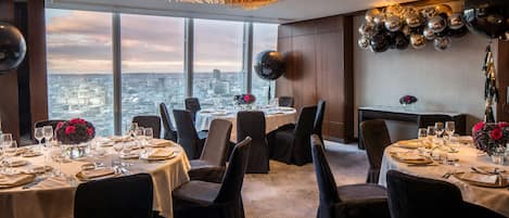 A dining area with tables set for a meal, large windows with a city view, and a chandelier.