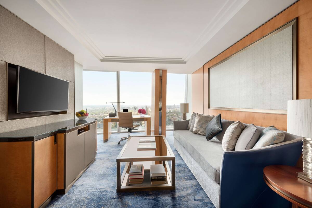 A modern hotel room with a flat-screen TV, a sofa, a coffee table, a desk, and a large window offering a city view.