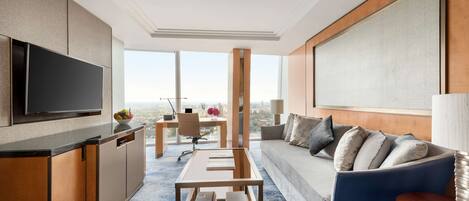 A modern hotel room with a flat-screen TV, a sofa, a coffee table, a desk, and a large window offering a city view.