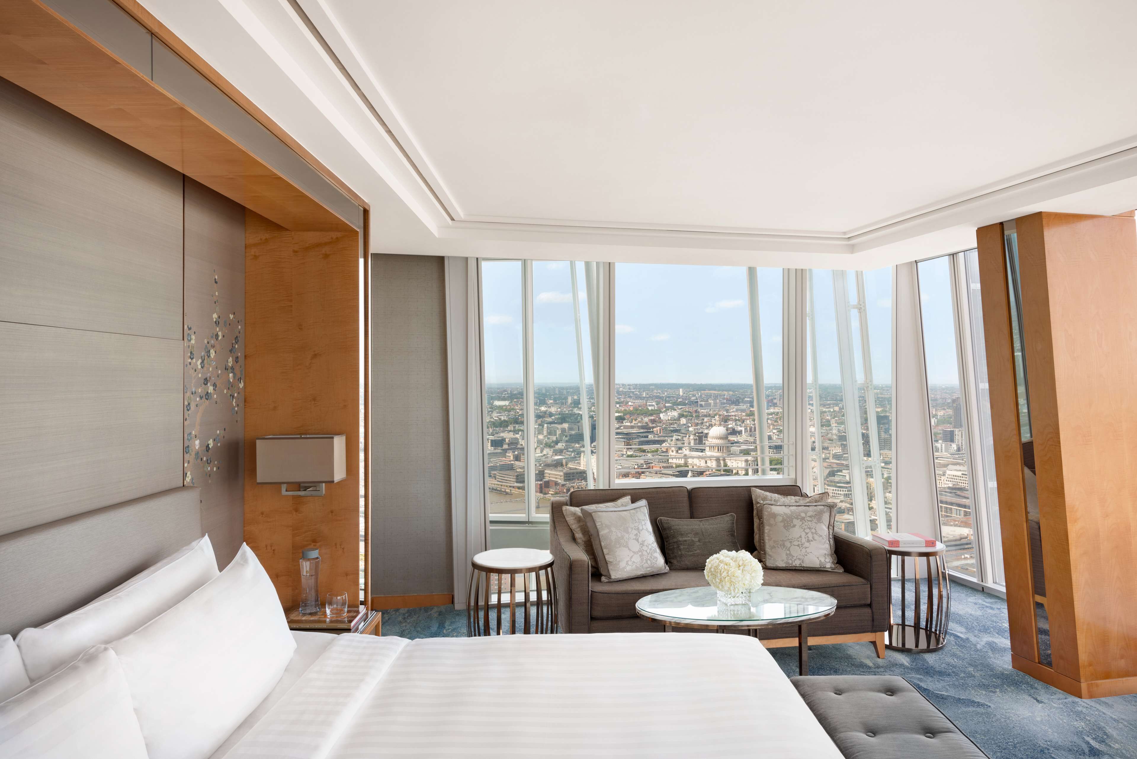 A modern hotel room with a large bed, a sofa, a small round table, and a view of the city through large windows.