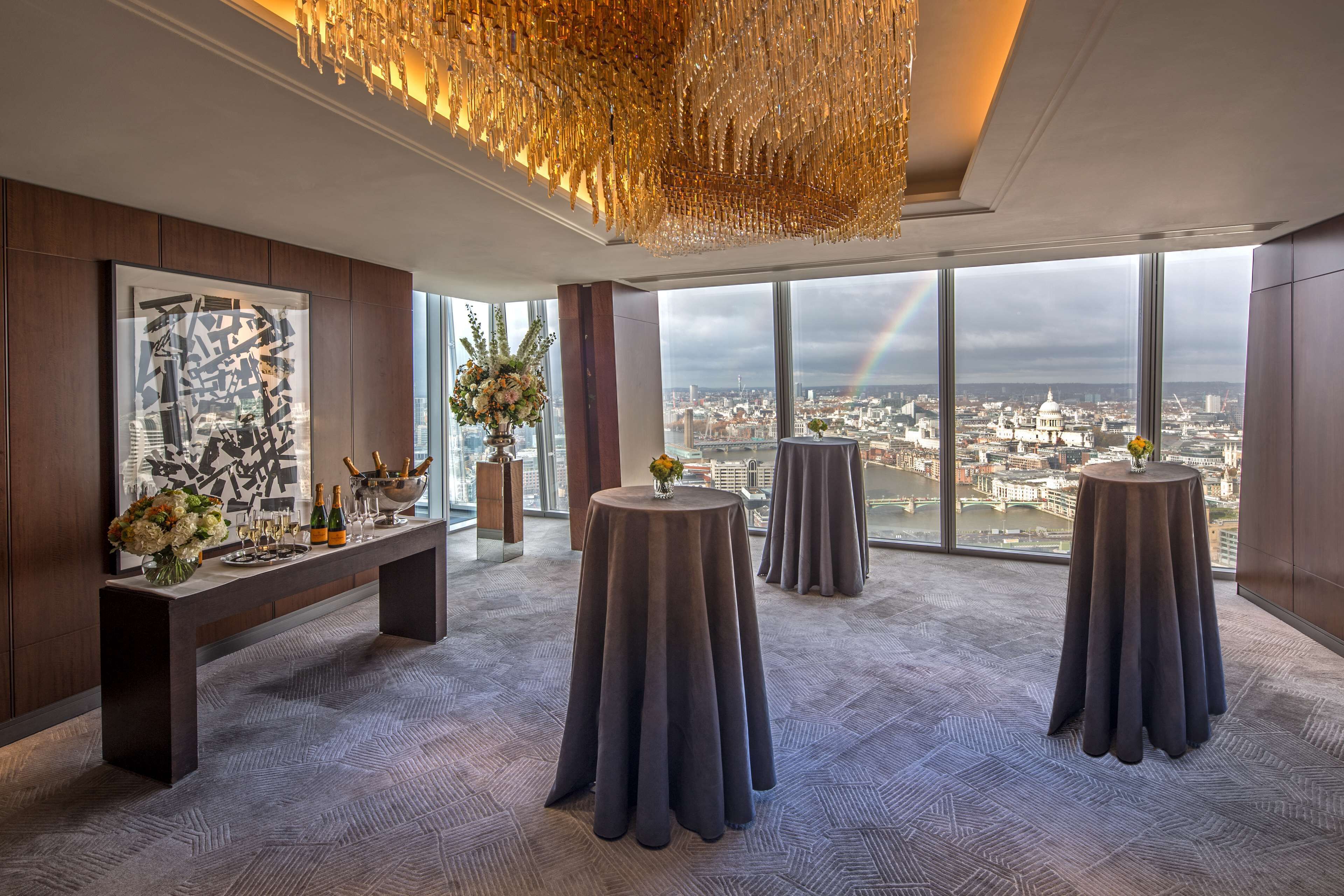 A high-rise hotel room with large windows, a city view, a chandelier, and a round table with a vase of flowers.
