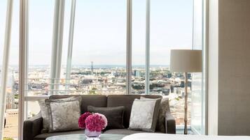 A hotel room with a large window offering a city view, a sofa with decorative pillows, and a small table with a vase.