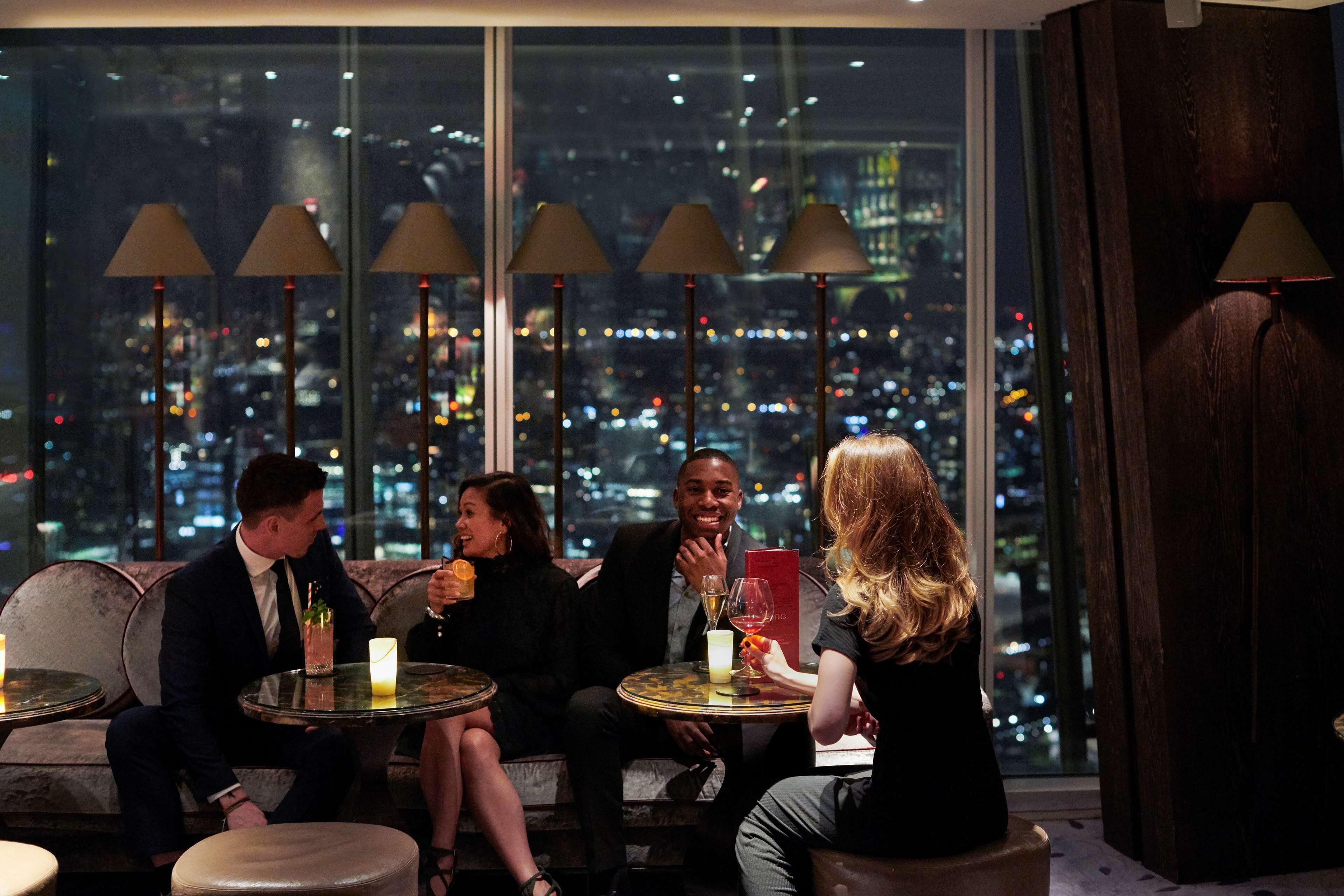A group of people seated at a table with a city view, in a room with large windows and multiple lamps.