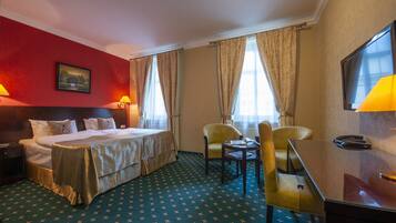 A hotel room with a bed, two chairs, a desk, a TV, and a painting on the wall.