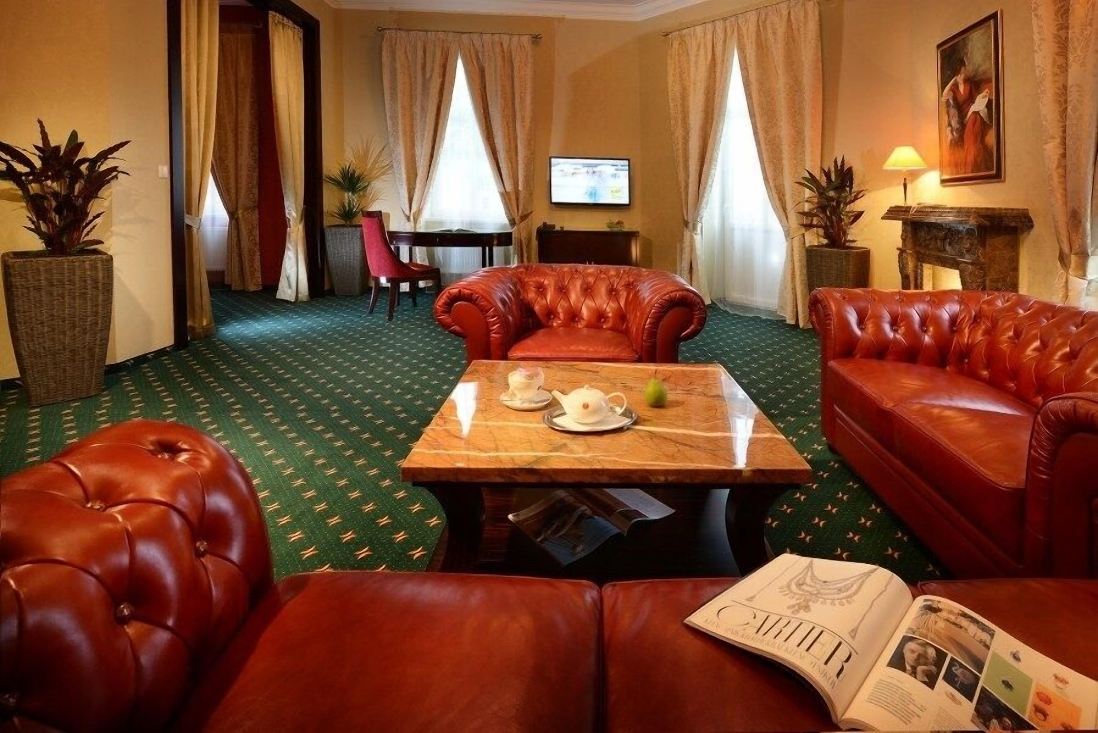 Habitación de hotel con alfombra verde estampada, sofás de cuero rojo, una mesa de centro de madera con una tetera y una pera, y un televisor en la pared.