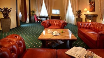 Habitación de hotel con alfombra verde estampada, sofás de cuero rojo, una mesa de centro de madera con una tetera y una pera, y un televisor en la pared.