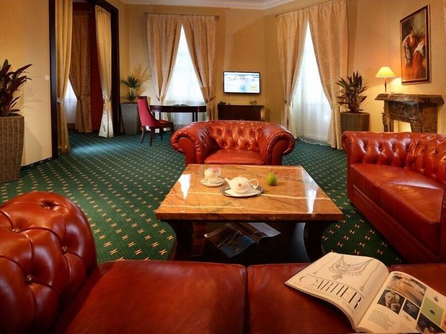 Habitación de hotel con alfombra verde estampada, sofás de cuero rojo, una mesa de centro de madera con una tetera y una pera, y un televisor en la pared.