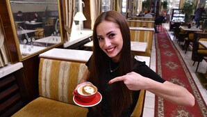 A person seated at a table with a cup of coffee, pointing at it.