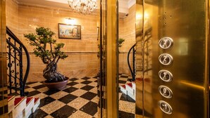An elevator interior with a checkered floor, a potted bonsai tree, a chandelier, and a painting on the wall.