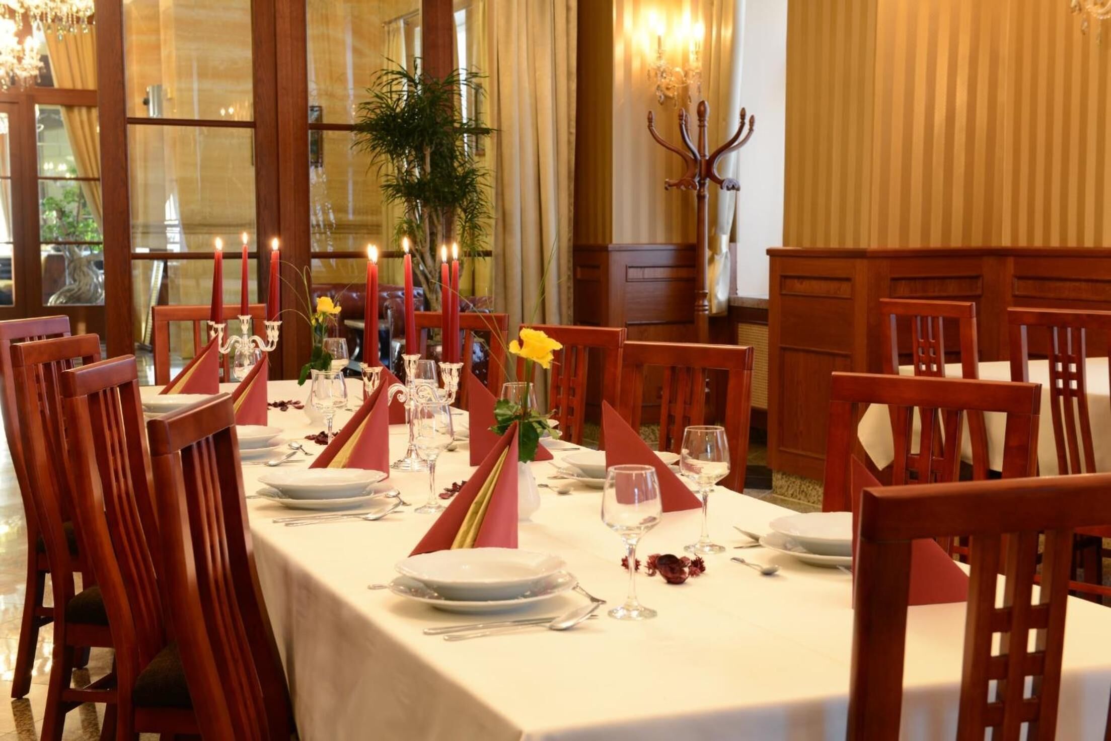 A dining table set for a meal with red napkins, candles, and a vase with yellow flowers.