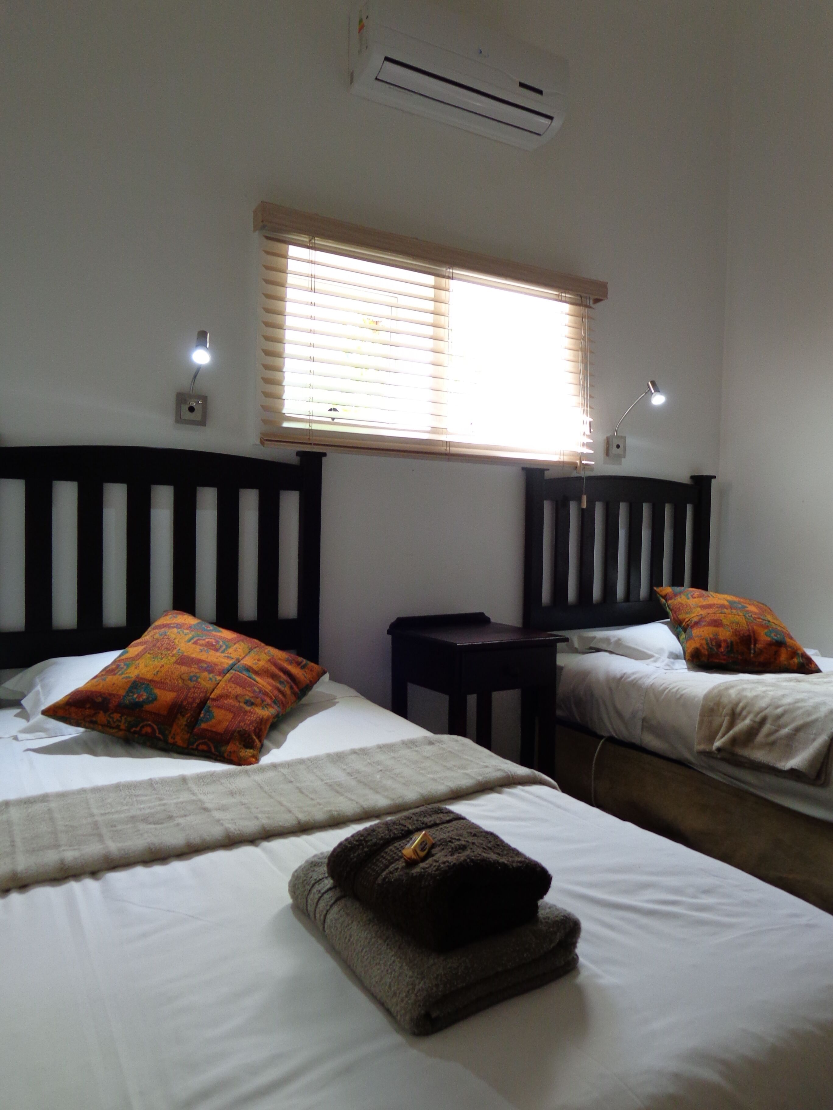 Habitación con dos camas, cada una con almohada y mesita de noche. Se observa una ventana con persianas, un aire acondicionado de pared y una lámpara en el techo.