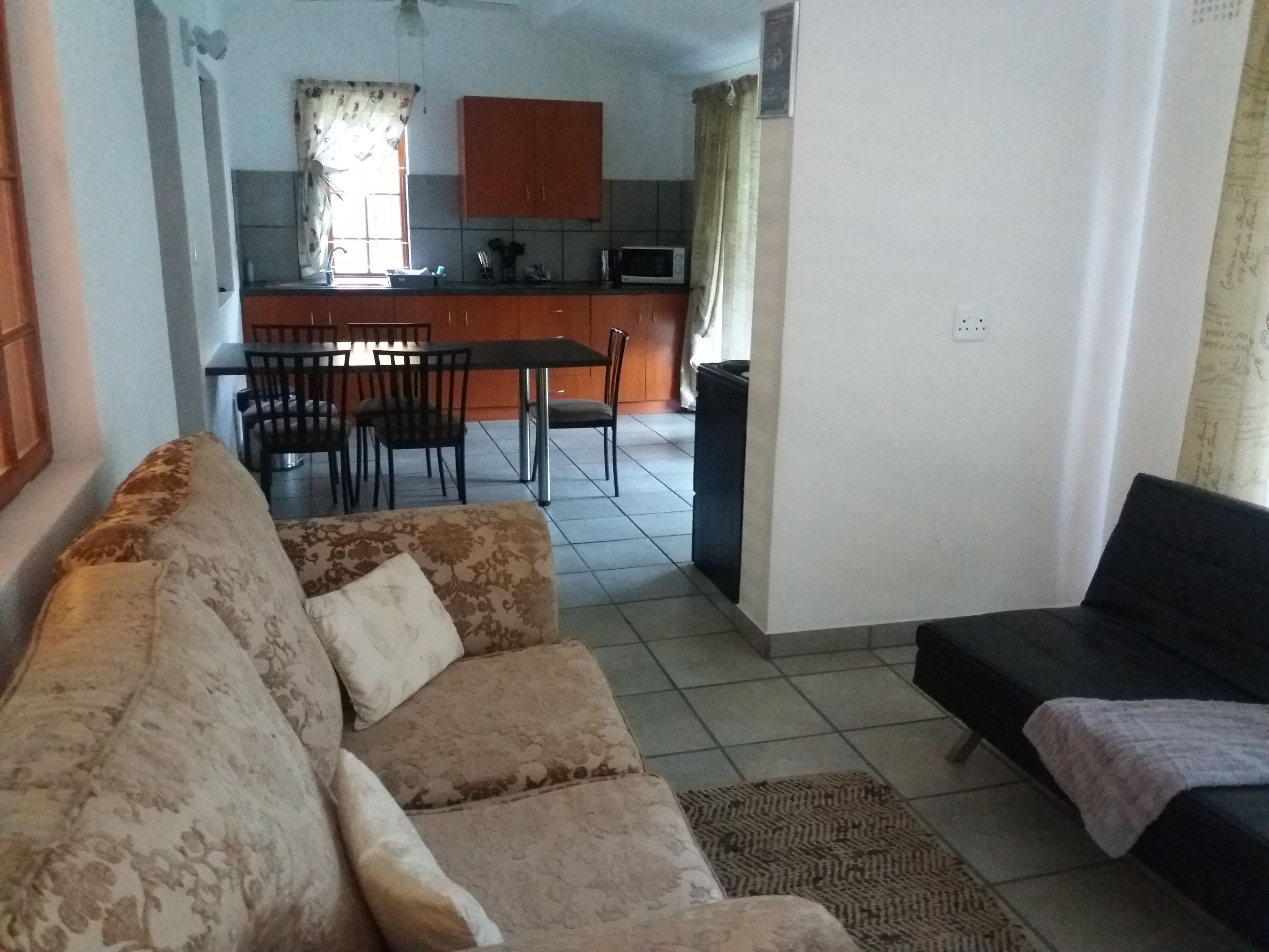 A living room with a sofa and a black couch, a dining area with chairs and a table, and a kitchen with cabinets and a microwave.