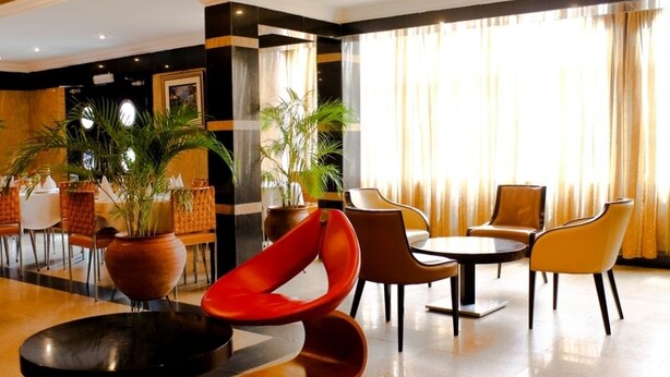 Un hall d’entrée moderne avec différents types de sièges, notamment un fauteuil rouge, des fauteuils marron et une table ronde. On y trouve également des plantes en pot et une grande fenêtre avec des rideaux.